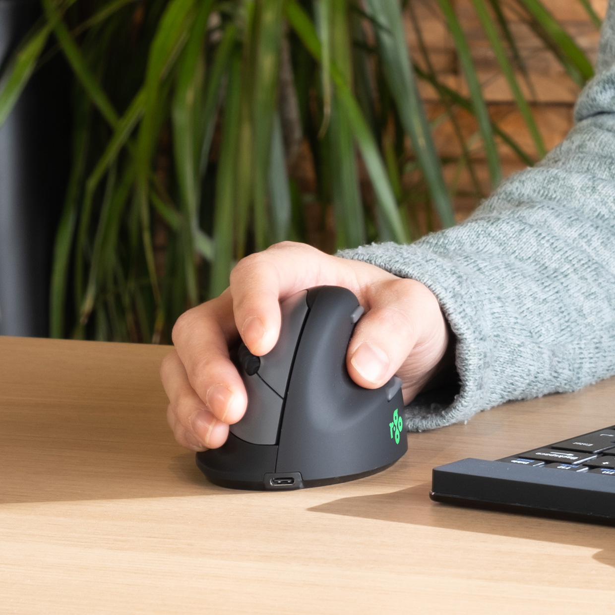 Die Hand einer Person benutzt eine schwarze ergonomische Maus auf einem Holztisch. Eine Tastatur ist im Hintergrund.