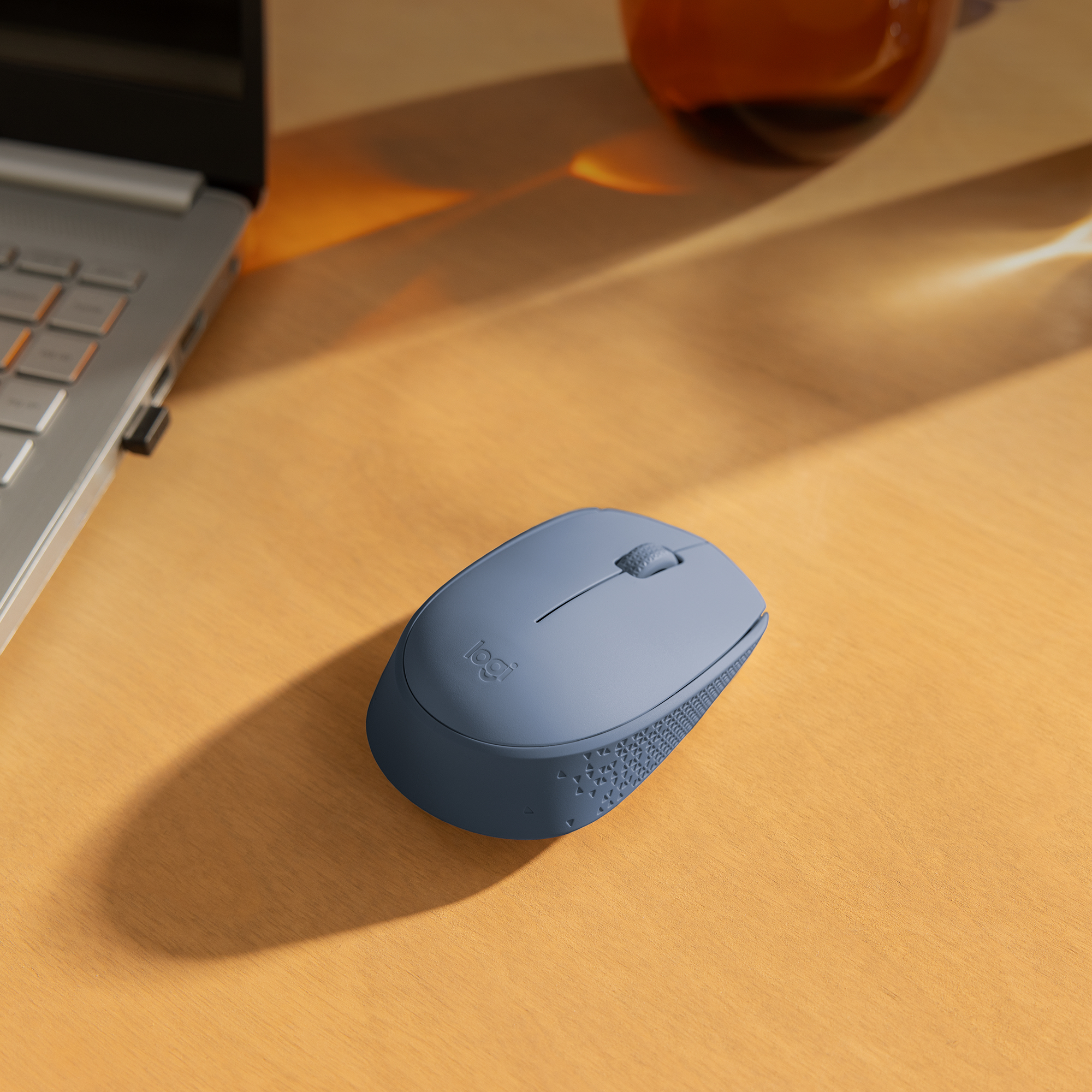 Blaue Maus auf einem Holztisch neben einem Laptop und einem Glas Flüssigkeit.