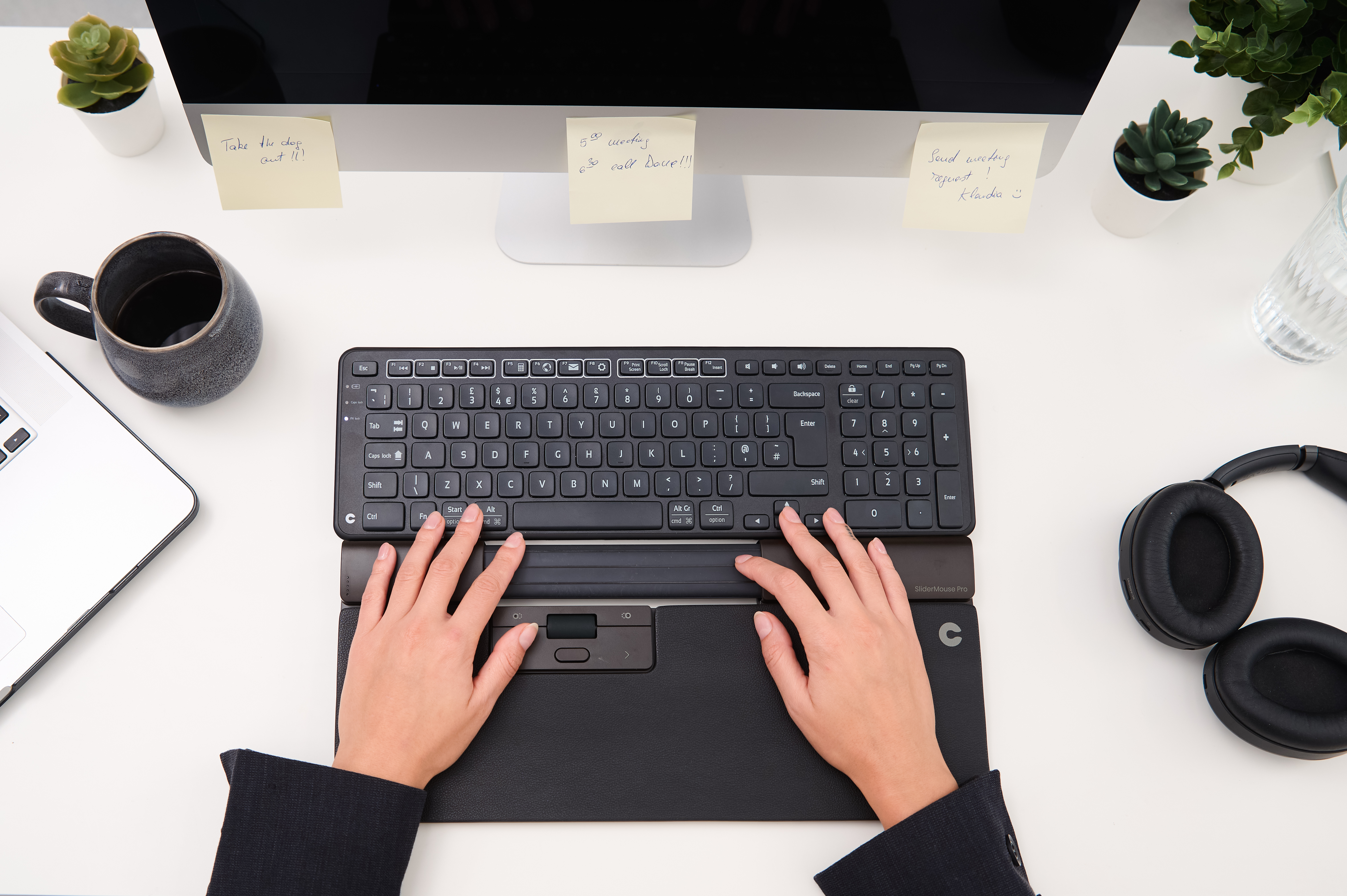 Mani che digitano su tastiera nera con poggiapolsi. Monitor, piante, tazza di caffè e accessori sulla scrivania.