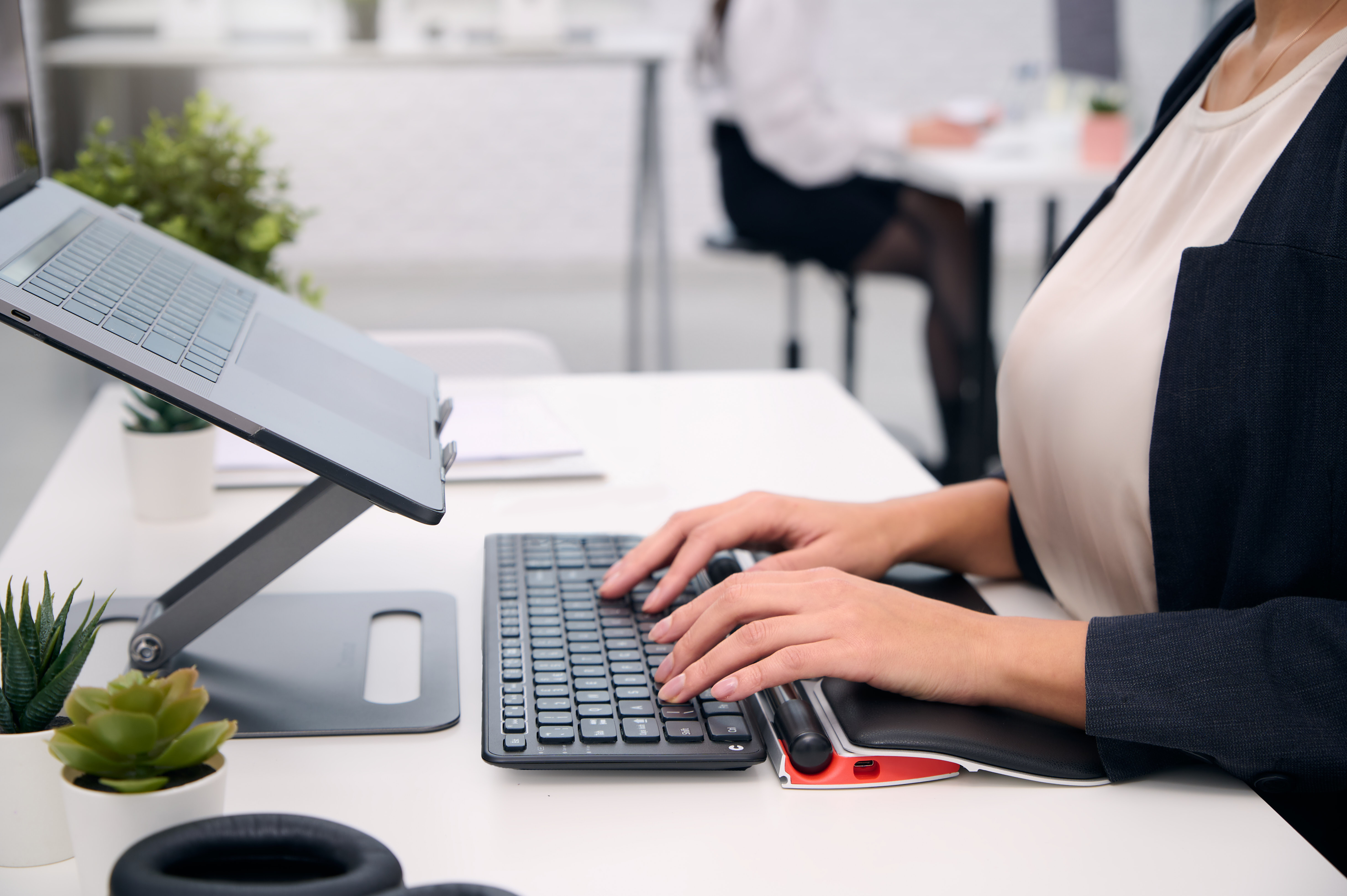 Osoba pisząca na klawiaturze z laptopem na stojaku. Biurko z roślinami i akcesoriami, druga osoba w tle.