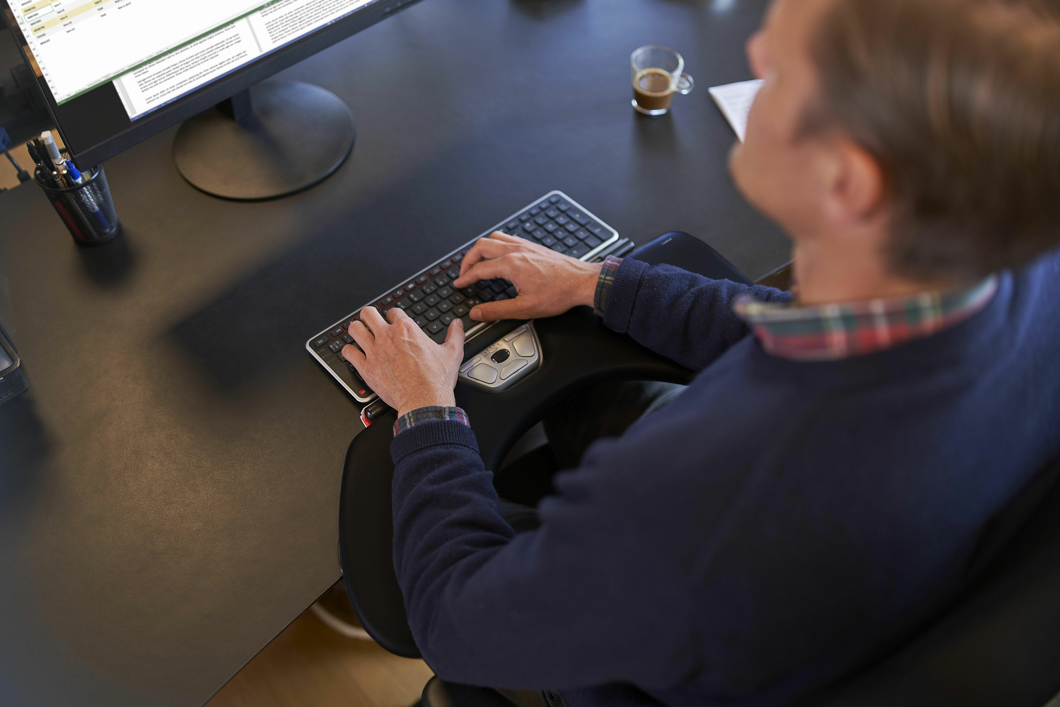 Mężczyzna używający ergonomicznej klawiatury i myszy przy biurku. Obecny monitor komputera i filiżanka kawy.