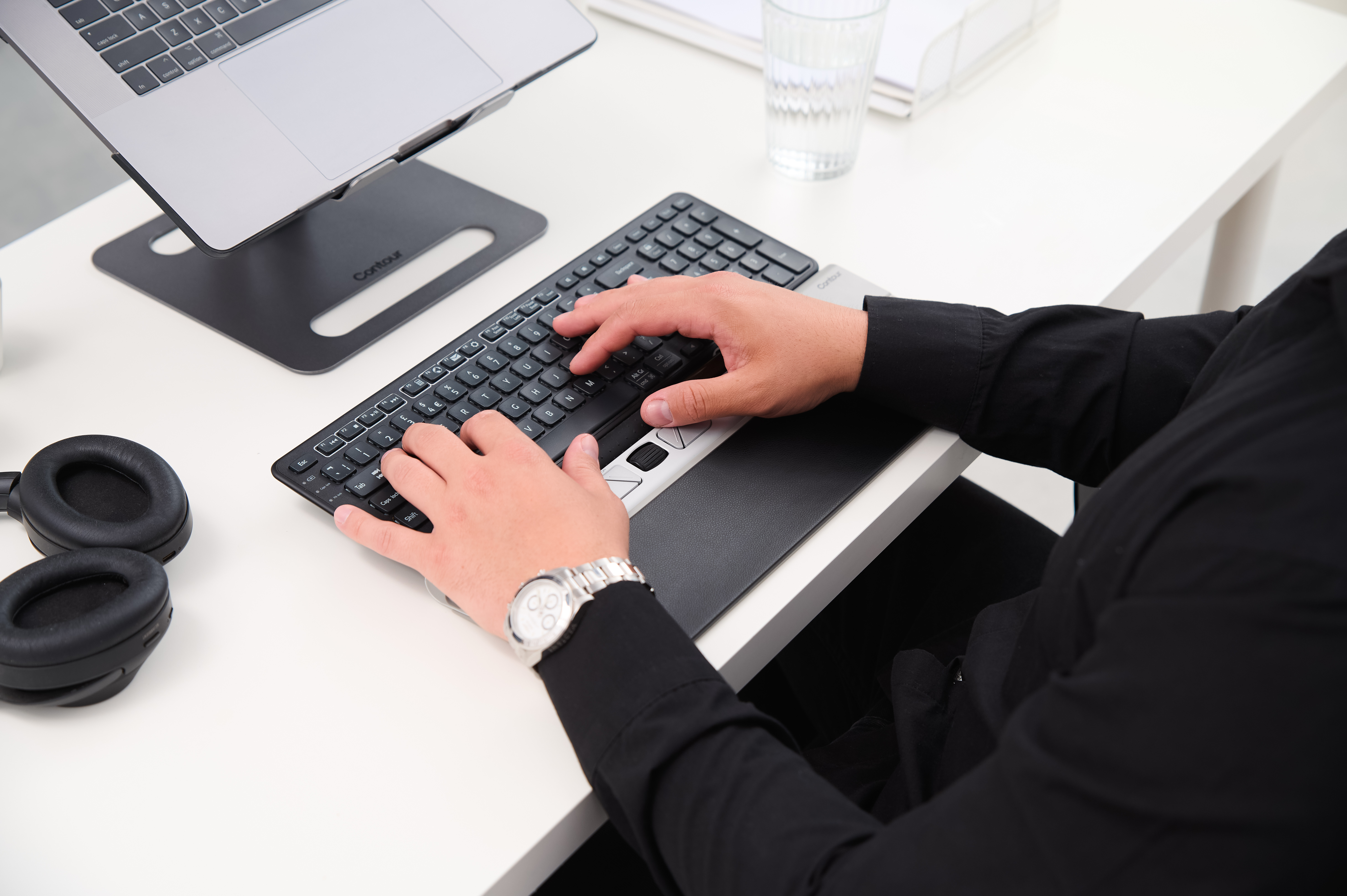 Osoba pisząca na klawiaturze z podpórką pod nadgarstek. Na biurku monitor, rośliny i słuchawki.