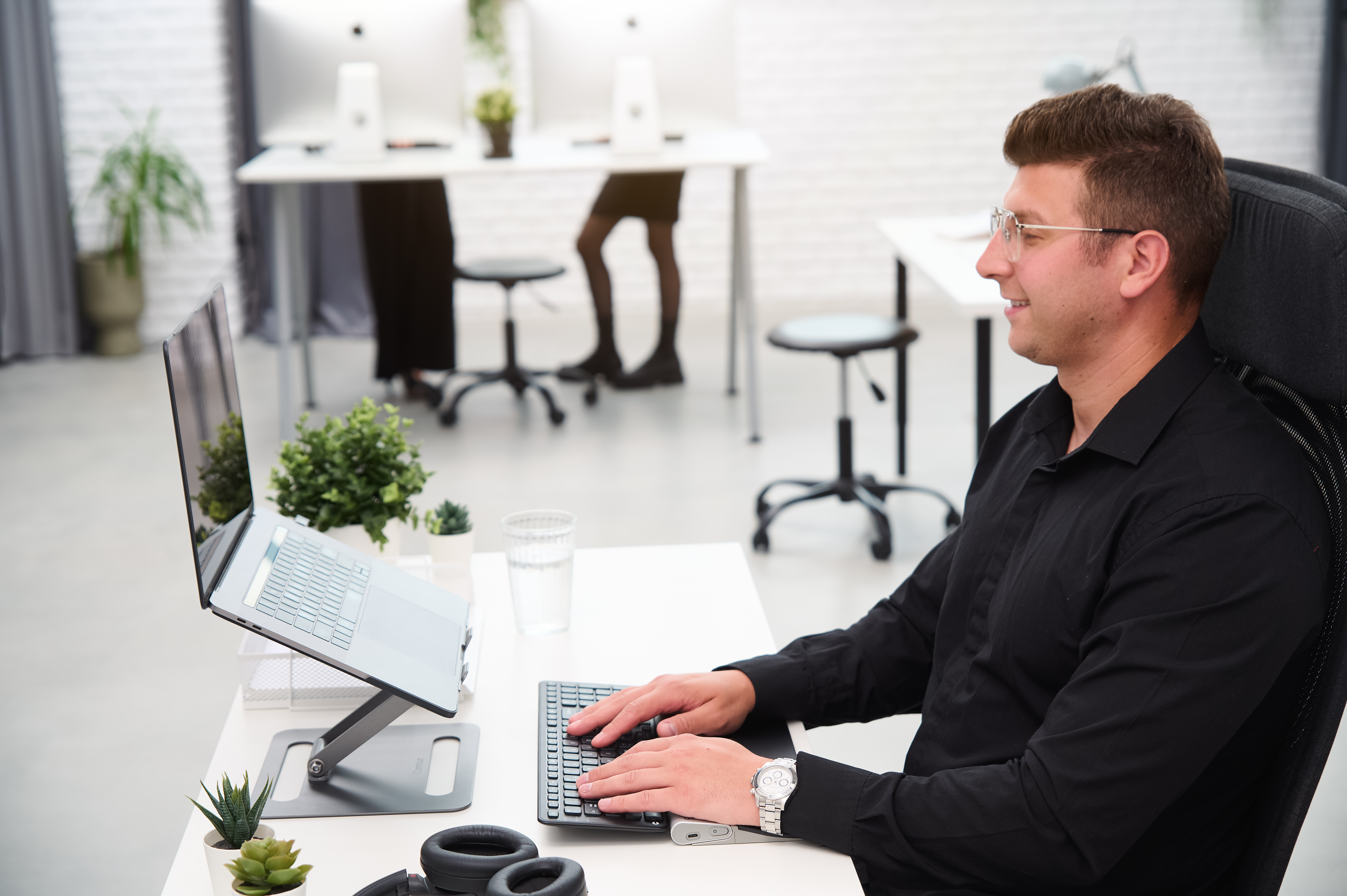 Mężczyzna używający laptopa z klawiaturą zewnętrzną. Biurko z roślinami i szklanką wody, białe tło.