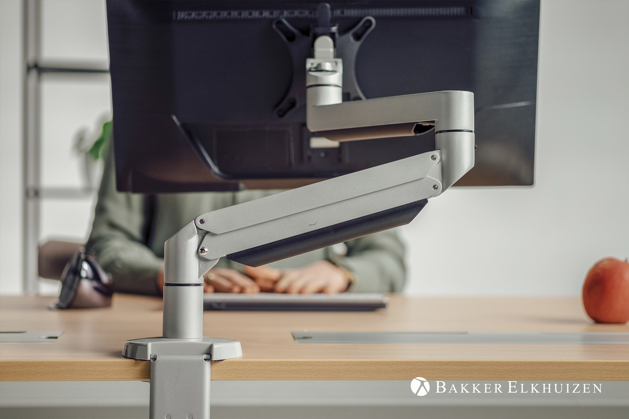 Monitor on a stand. The stand is silver. There is a desk and an apple in the background.