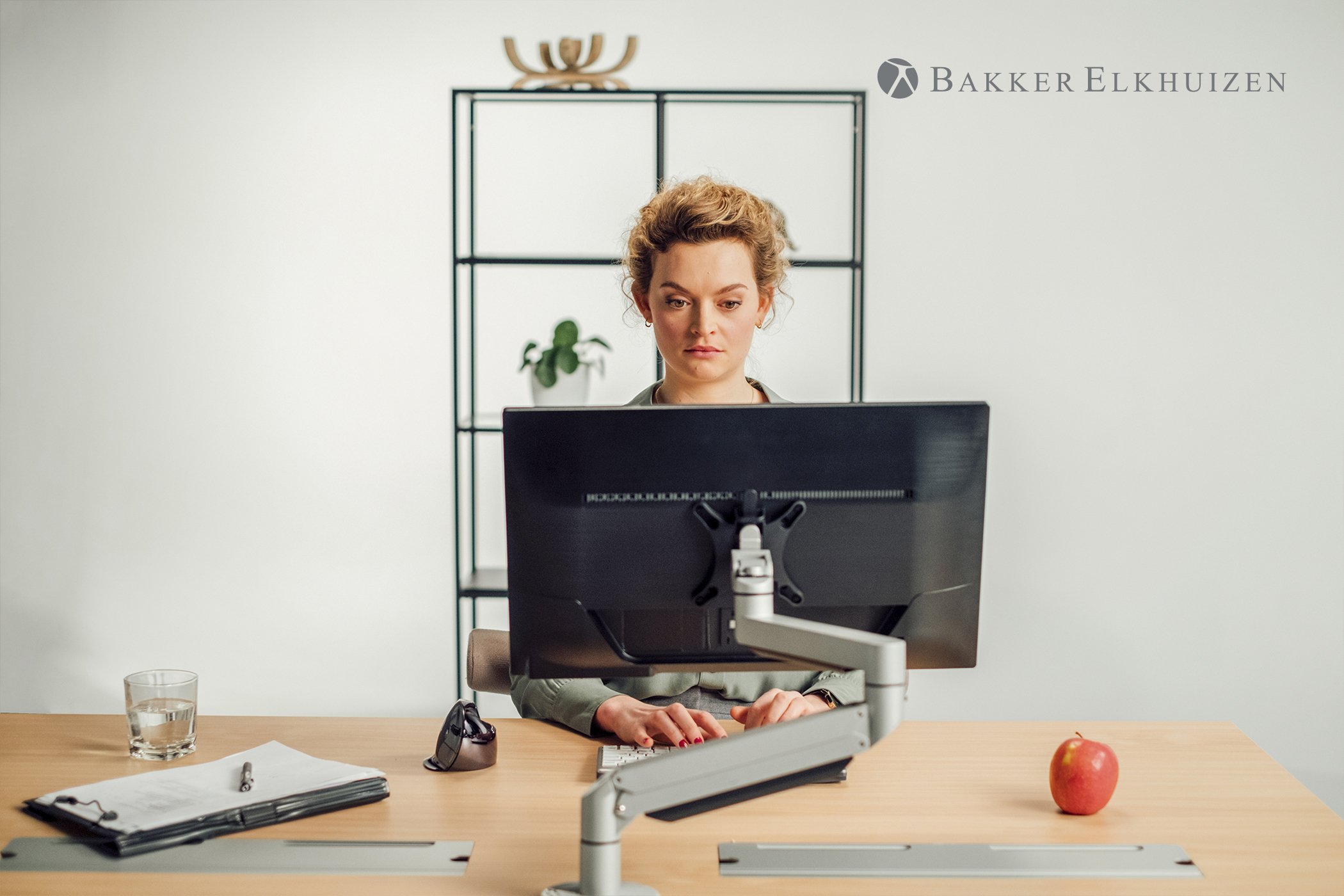 A woman is sitting at a desk using a computer. There is a glass of water, a notepad, and an apple.