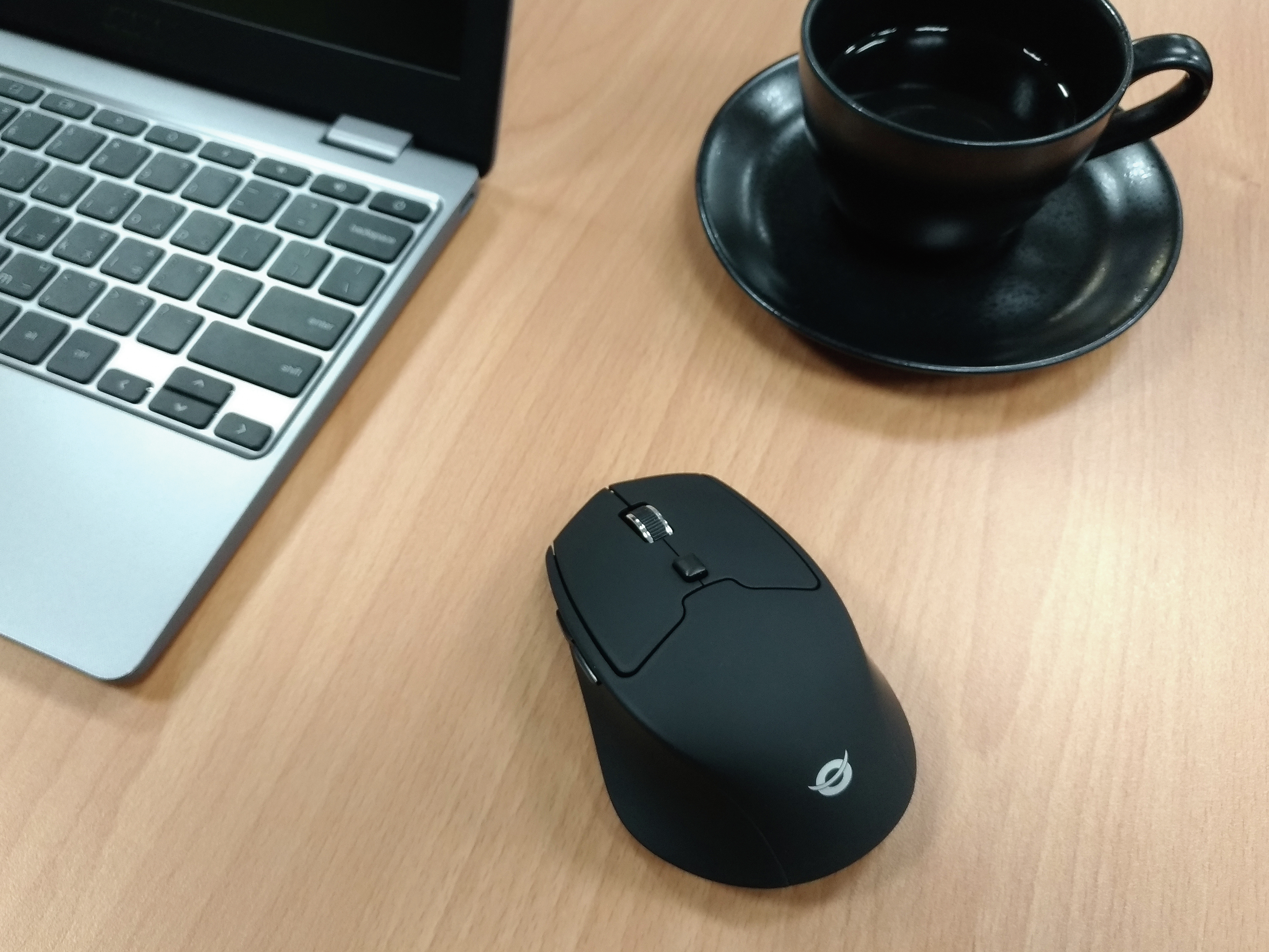 Een zwarte muis ligt op een houten bureau naast een laptop en een kopje koffie op een zwart bord.