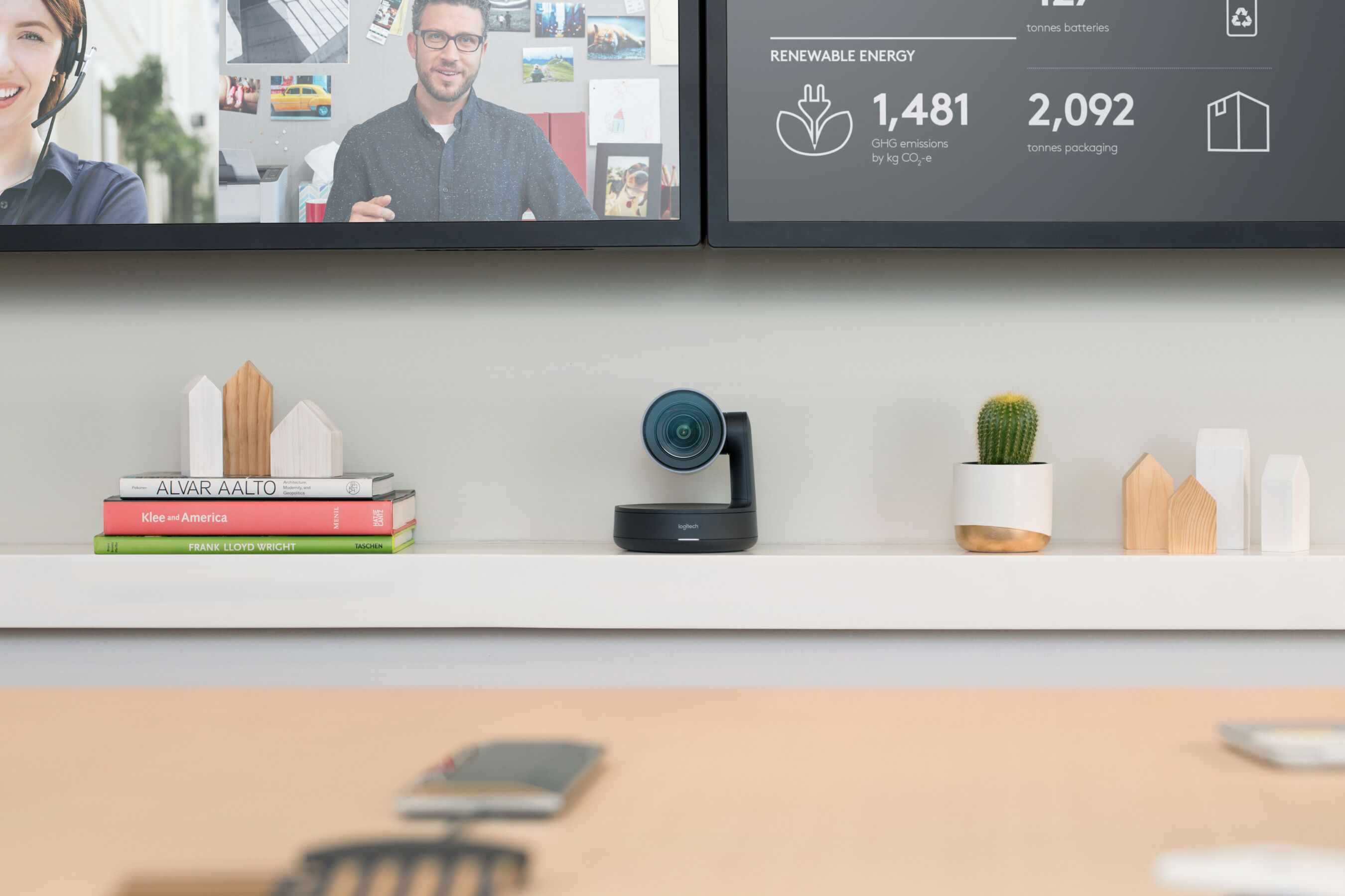 Eine Logitech-Webcam auf einem Regal mit Büchern, einem Kaktus und Holzhausdekorationen, mit verschwommenem Hintergrund.
