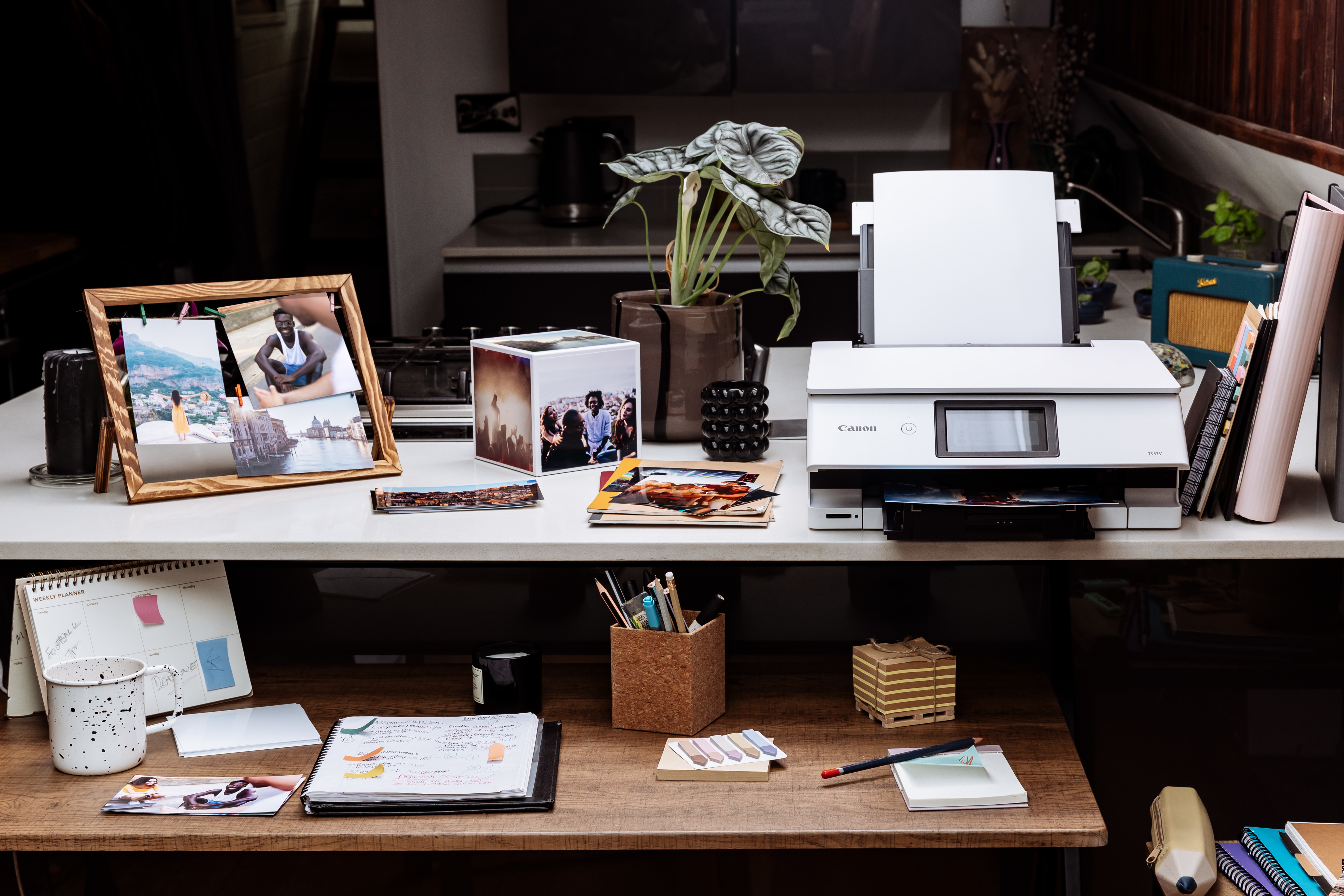 Biurko z drukarką, zdjęciami i artykułami piśmienniczymi. Roślina na biurku. Tło: kuchnia.