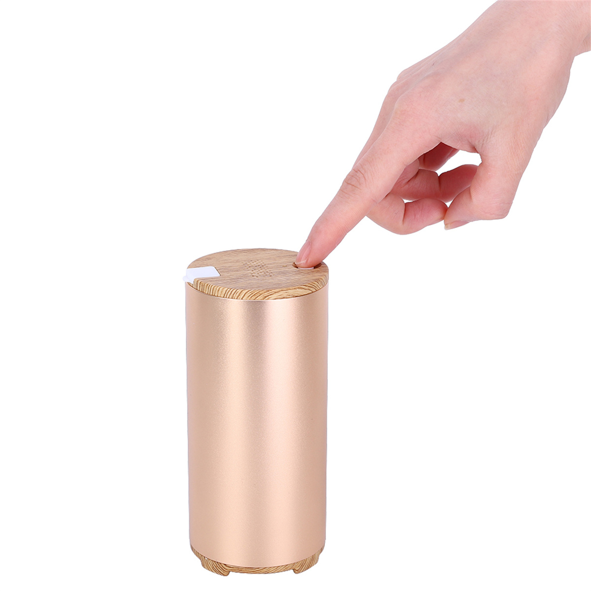 Ein gold- und holzfarbener Zylinder. Ein Finger drückt einen Knopf oben. Das Gerät steht auf weißer Oberfläche.