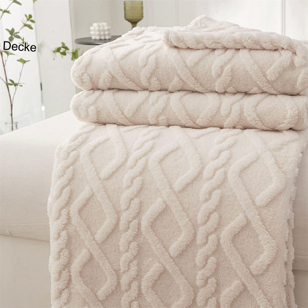 Gefaltete beige gemusterte Decken auf einem Bett, mit einem Blick auf eine Vase und Pflanze.