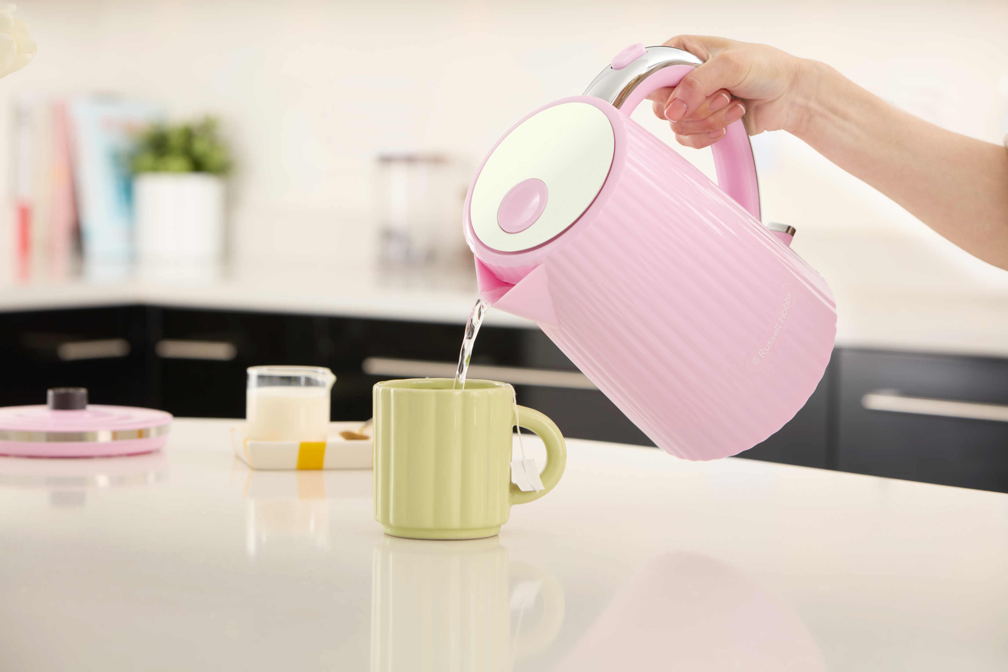 Una persona versa acqua da un bollitore rosa in una tazza verde, una lattiera e del cibo sul tavolo.