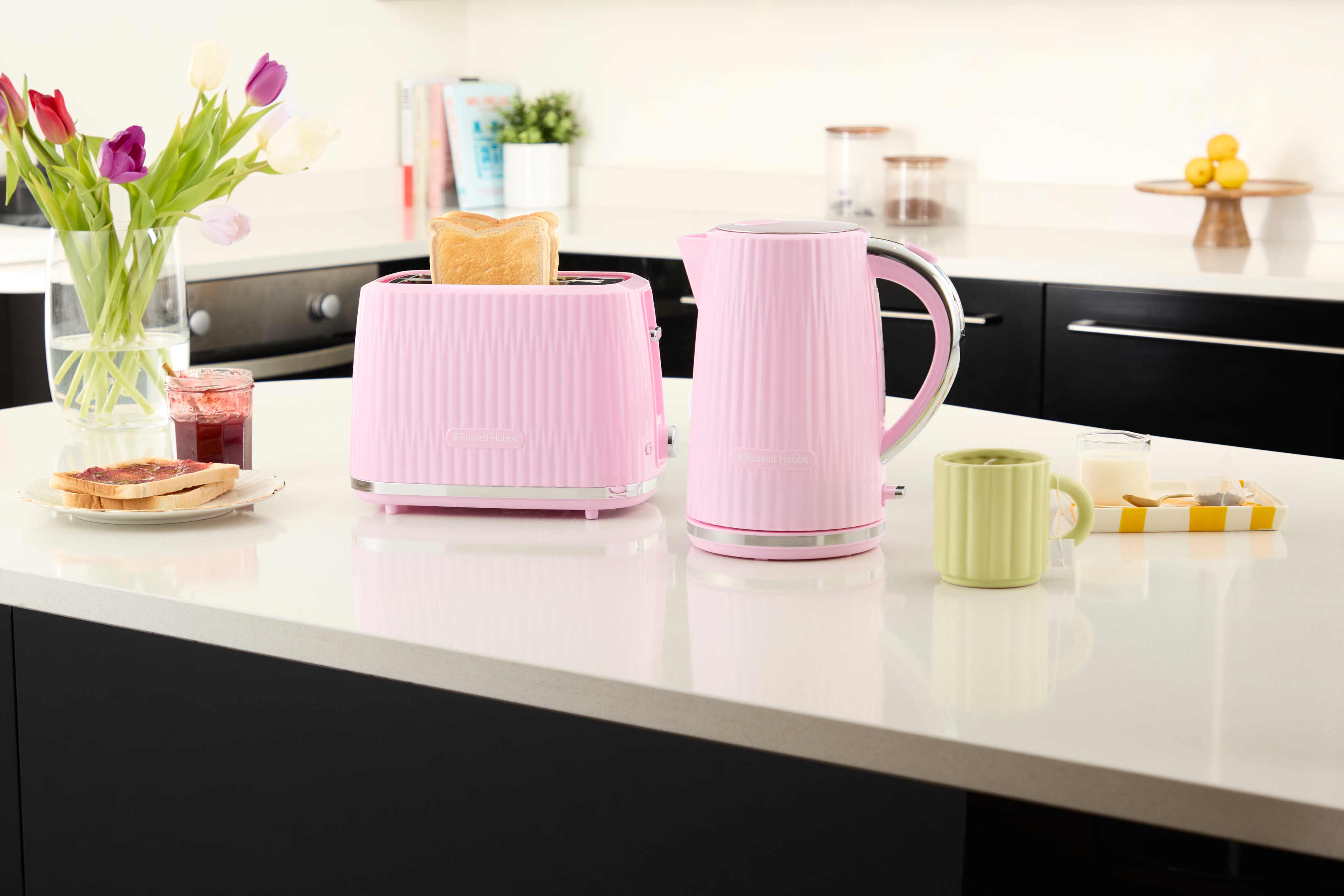 Set tostapane e bollitore rosa su piano bianco. Toast, fiori e tazza presenti.