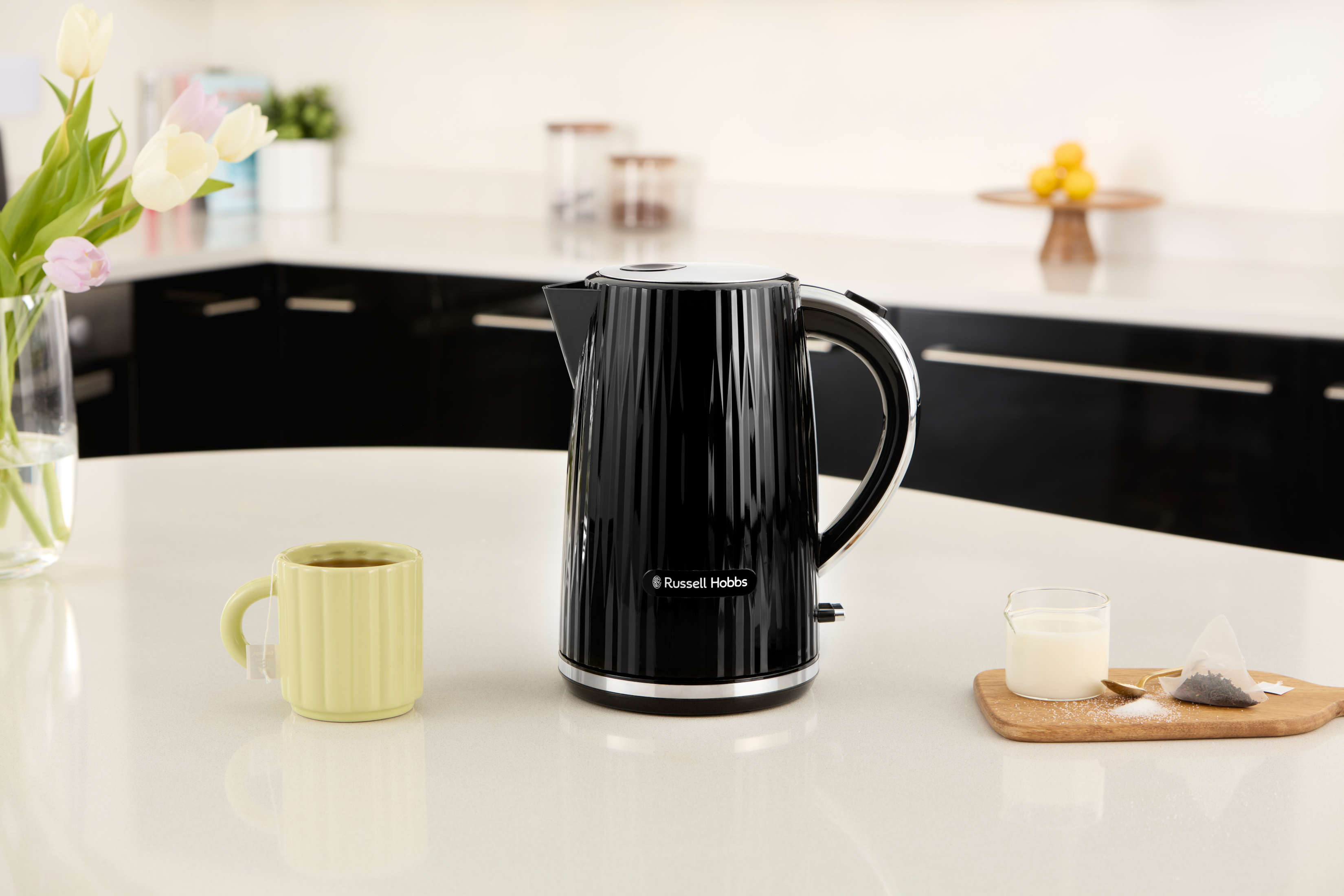 Una tetera eléctrica negra con una taza y flores en una mesa blanca. Hay una tabla de madera con bolsitas de té.