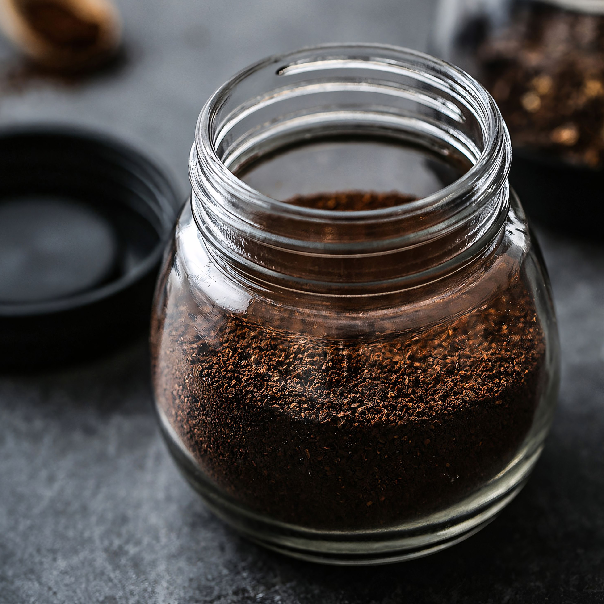 Un barattolo di vetro pieno di caffè macinato marrone scuro, senza coperchio, su una superficie scura.