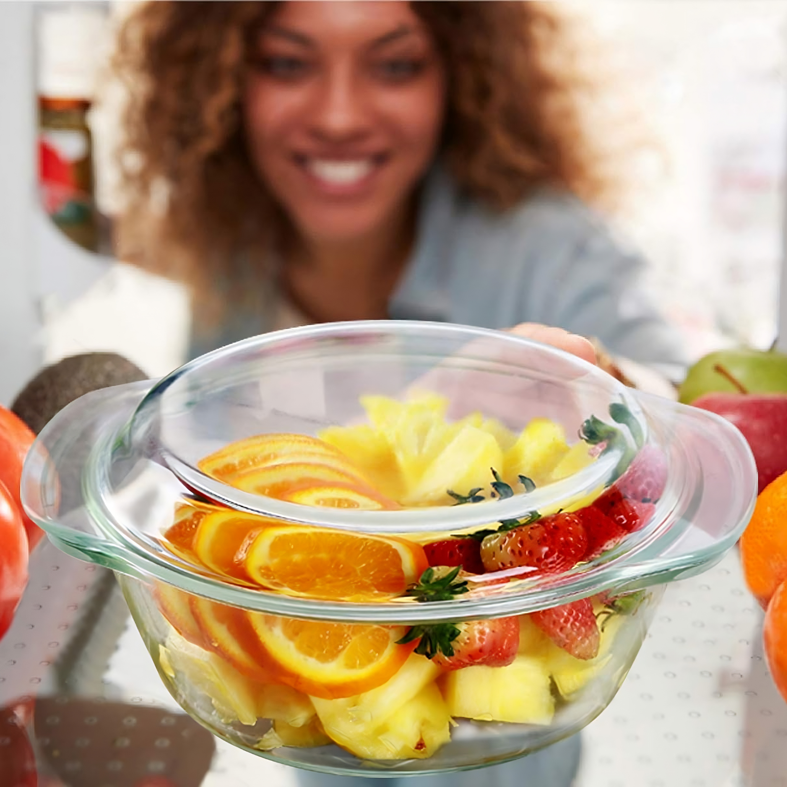 Eine Glasschüssel mit Obst gefüllt. Eine Frau greift danach und lächelt. Im Hintergrund gibt es mehr Früchte.