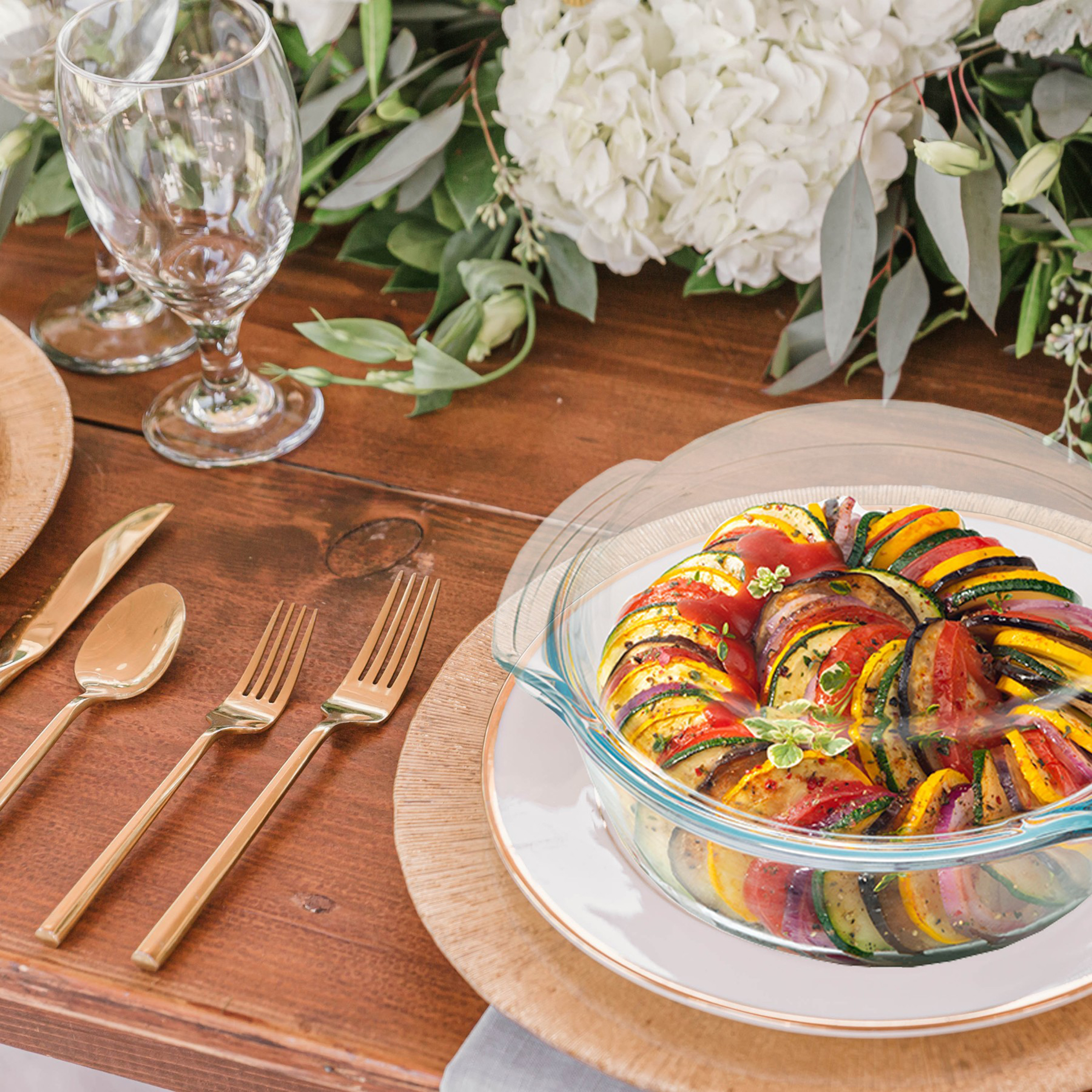 Tisch für Mahlzeit gedeckt, mit Ratatouille in Glasschale, goldenem Besteck und Gläsern.