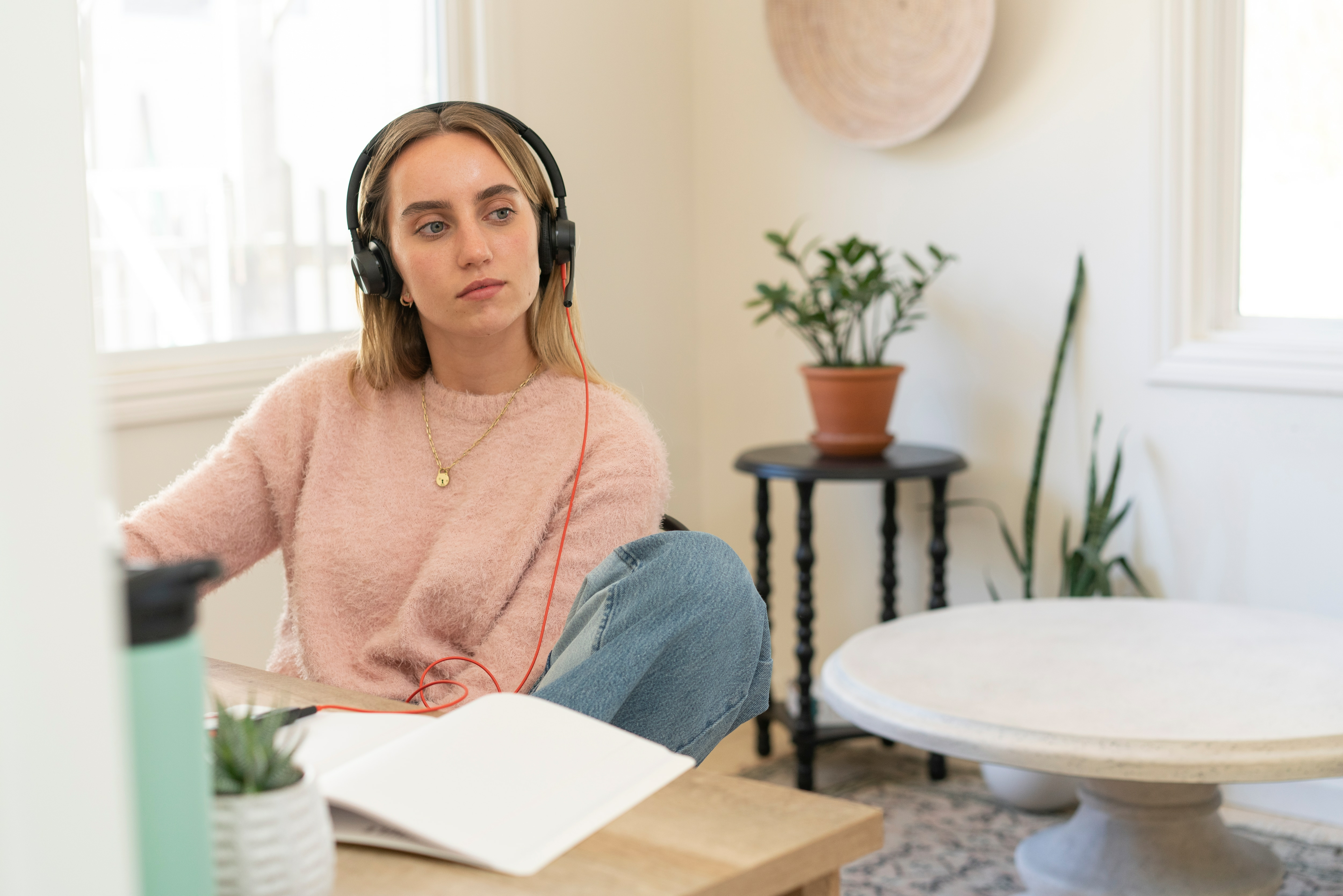 Kobieta w różowym swetrze i słuchawkach, pracująca w pokoju z oknem i stołem.