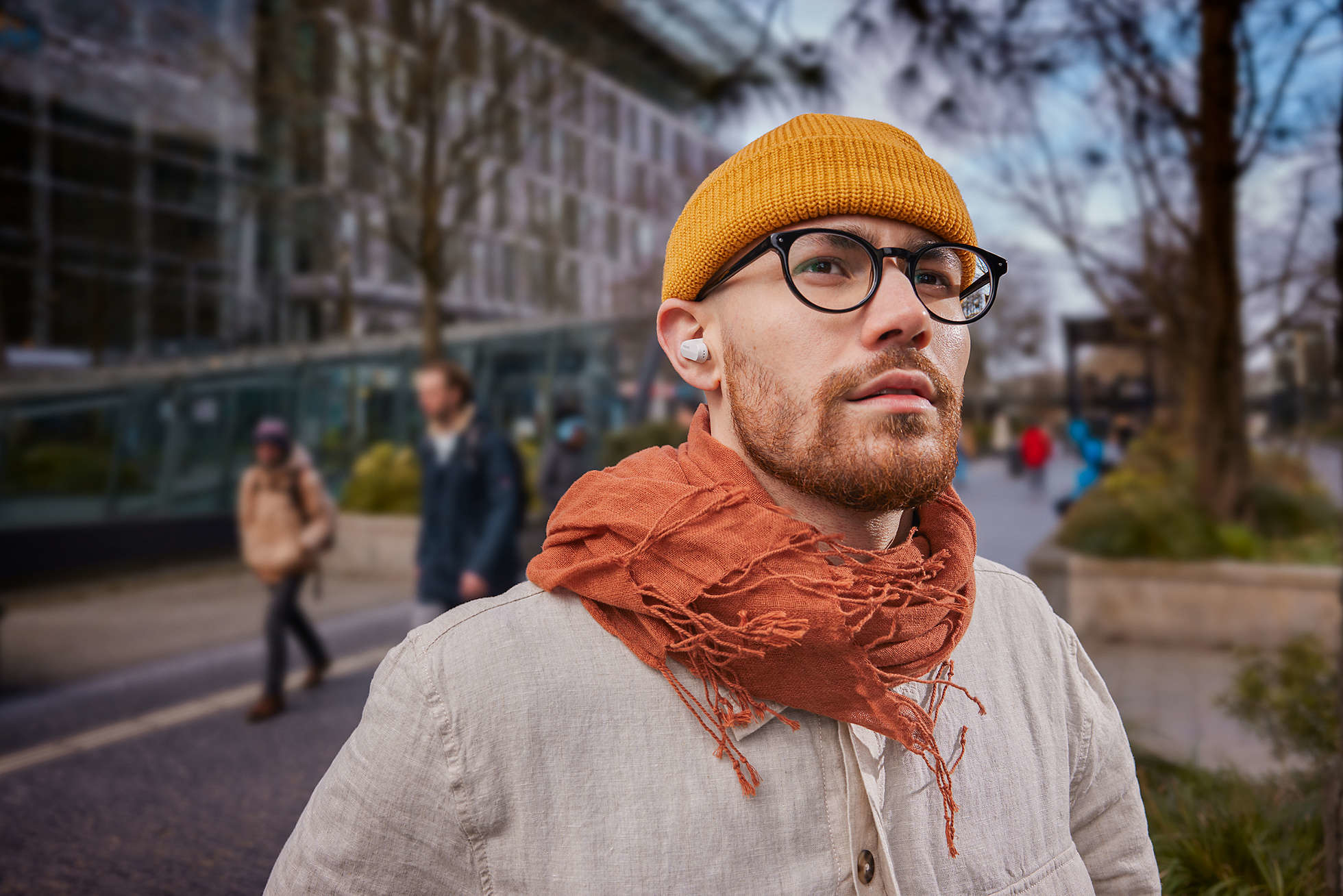 Ein Mann mit Brille, gelbem Hut und orangefarbenem Schal, mit Ohrhörern in den Ohren.