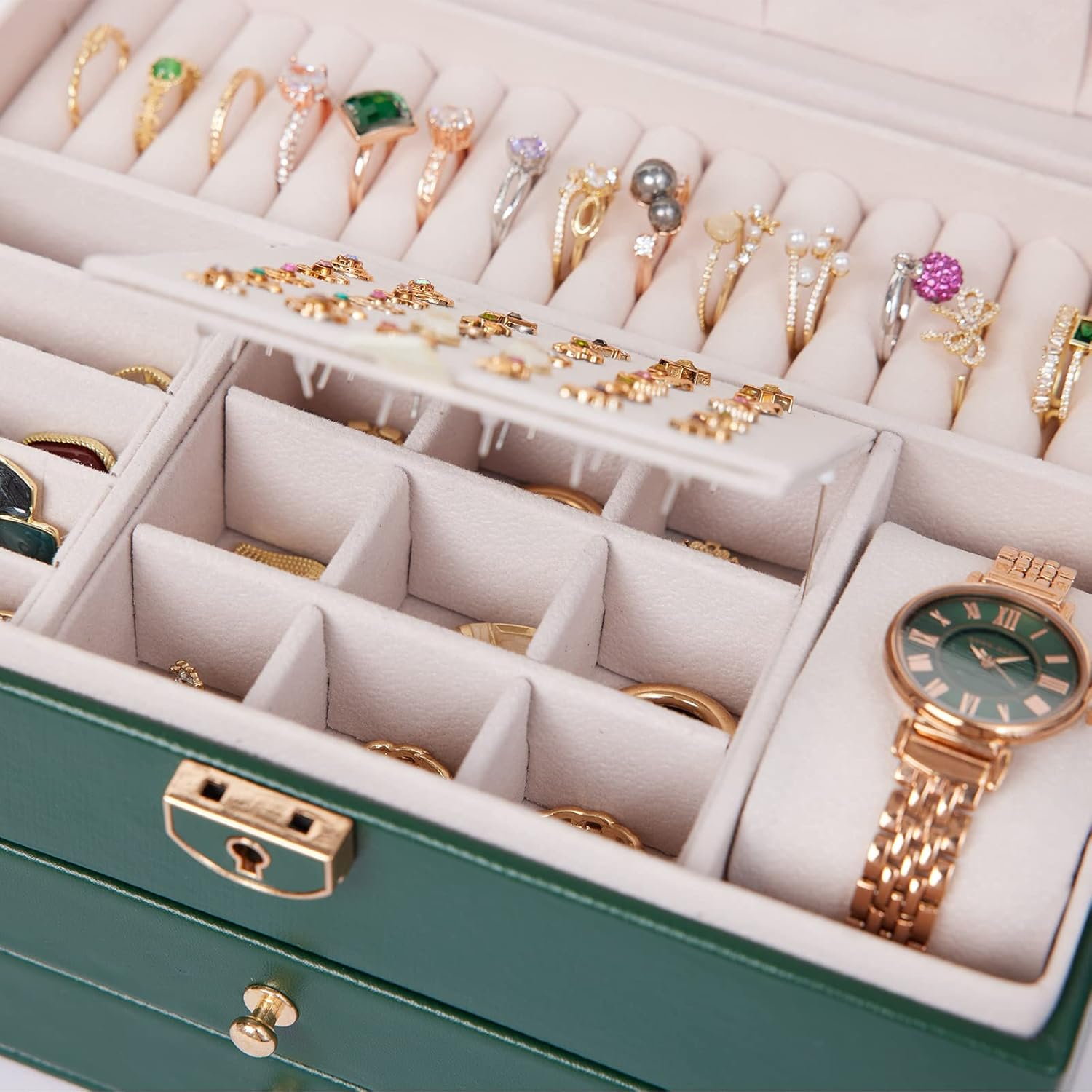 Offene grüne Schmuckschatulle mit Ringen, Uhr und Fächern auf weißer Oberfläche.