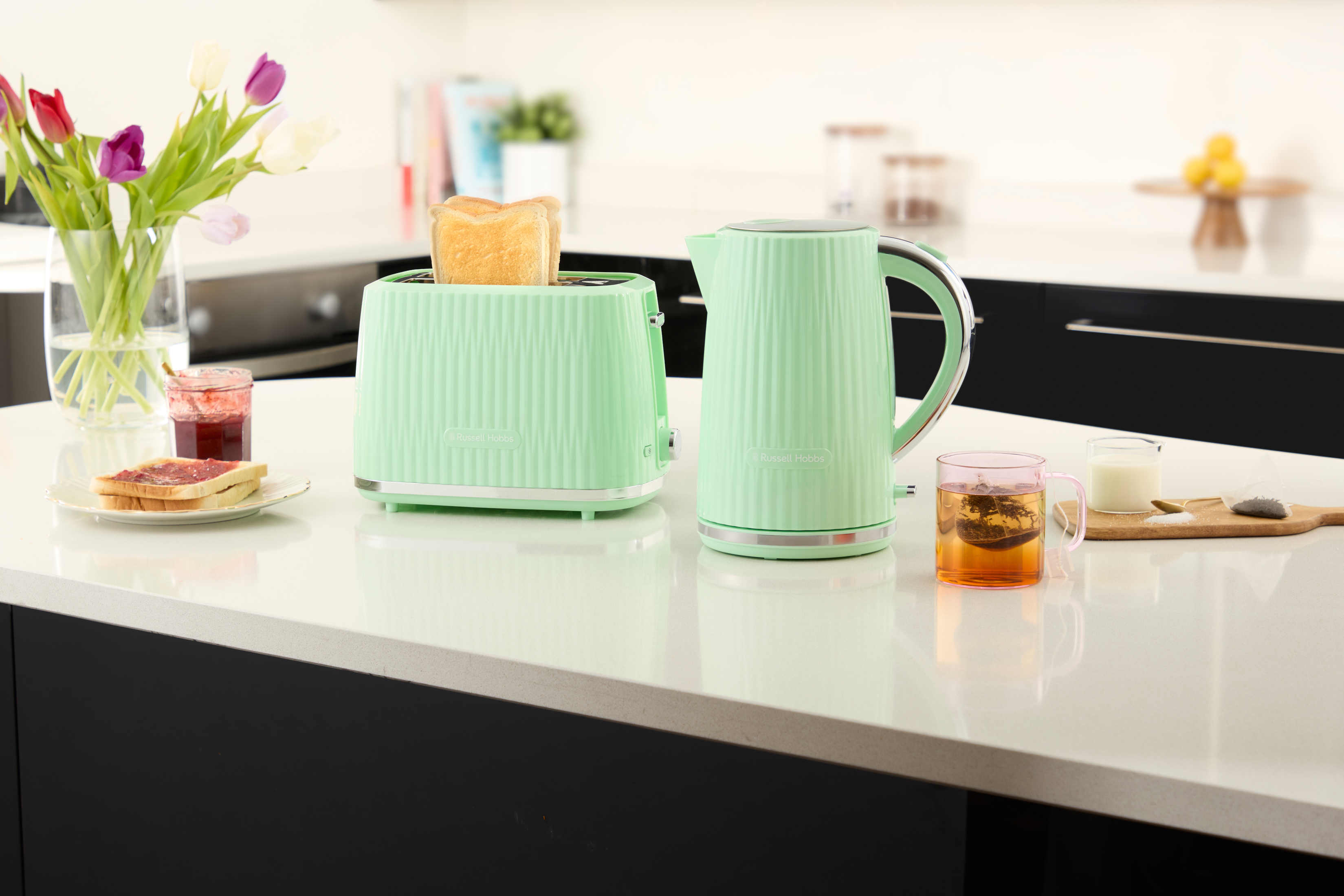 Tostapane e bollitore verde menta, con toast, marmellata, tè e latte su un bancone bianco.
