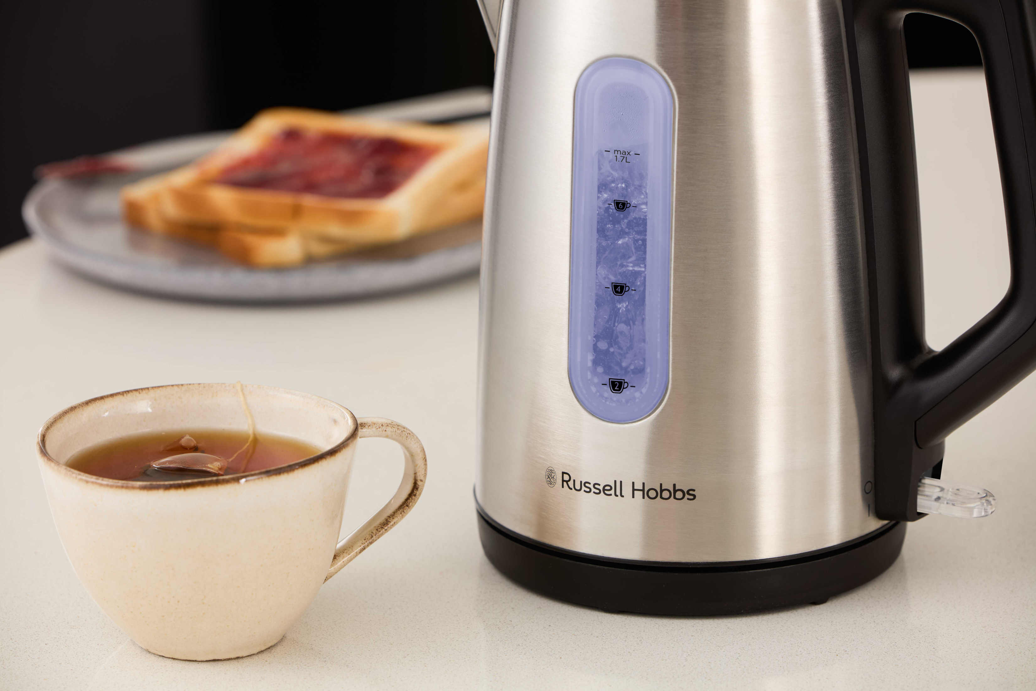 Ein elektrischer Wasserkocher aus Edelstahl mit blauer Wasserstandsanzeige neben einer Tasse Tee und Toast.