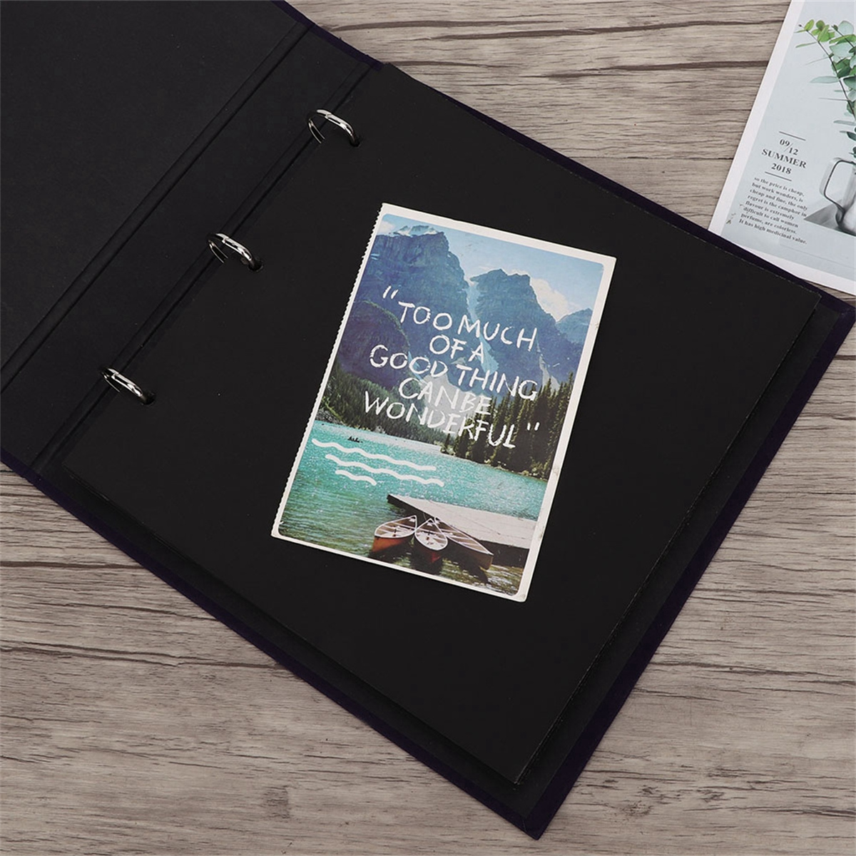 Offenes Fotoalbum mit Postkarte, Berge und See zeigend. Schwarze Seiten, Holzuntergrund sichtbar.
