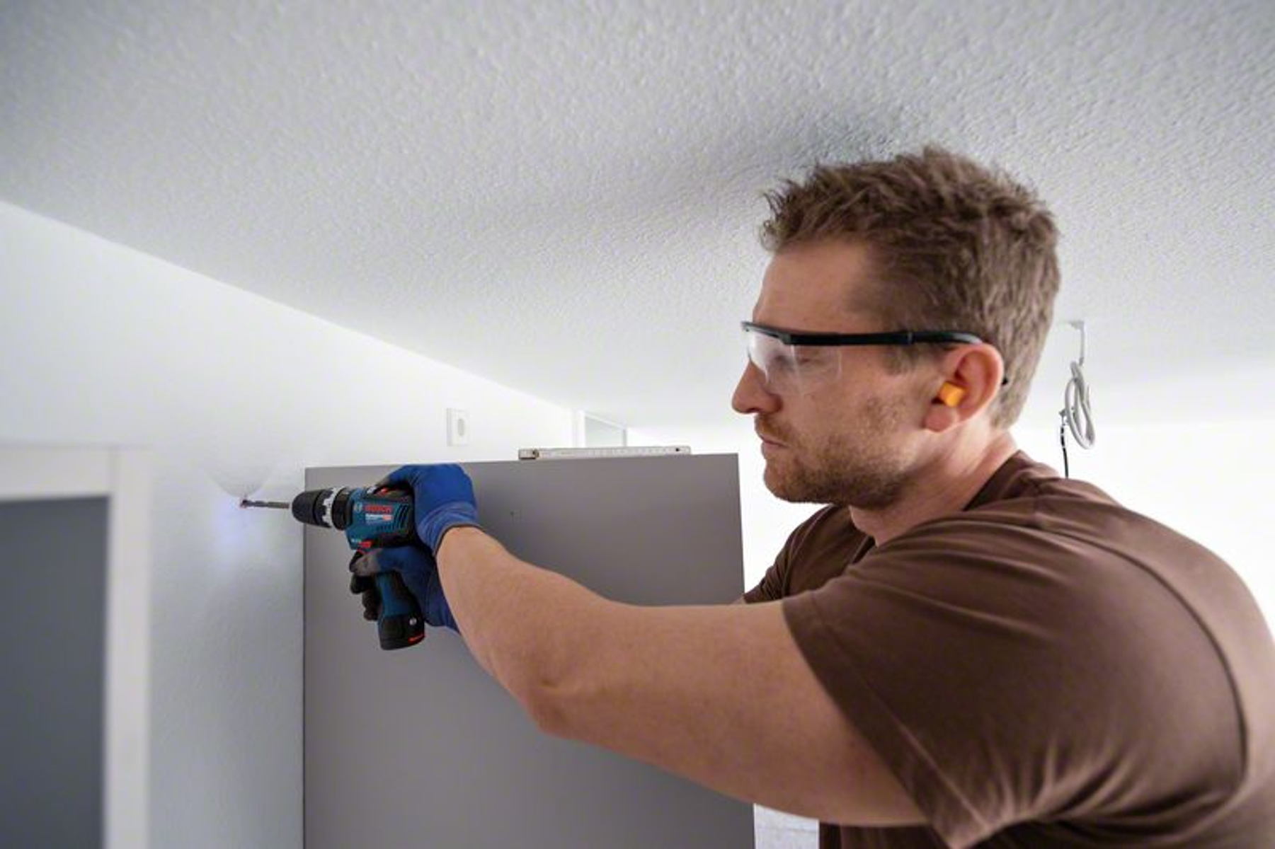 Mężczyzna wierci w ścianie wiertarką, nosząc okulary ochronne i zatyczki do uszu.