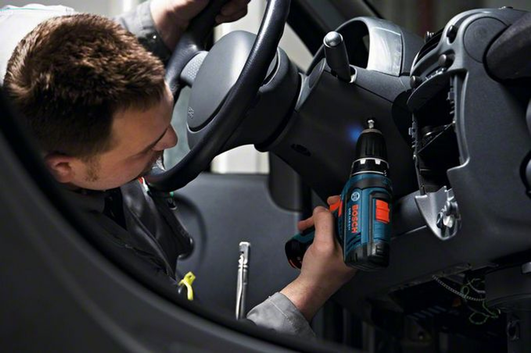 Mechanik używający wiertarki do pracy nad deską rozdzielczą samochodu, widoczna kierownica.