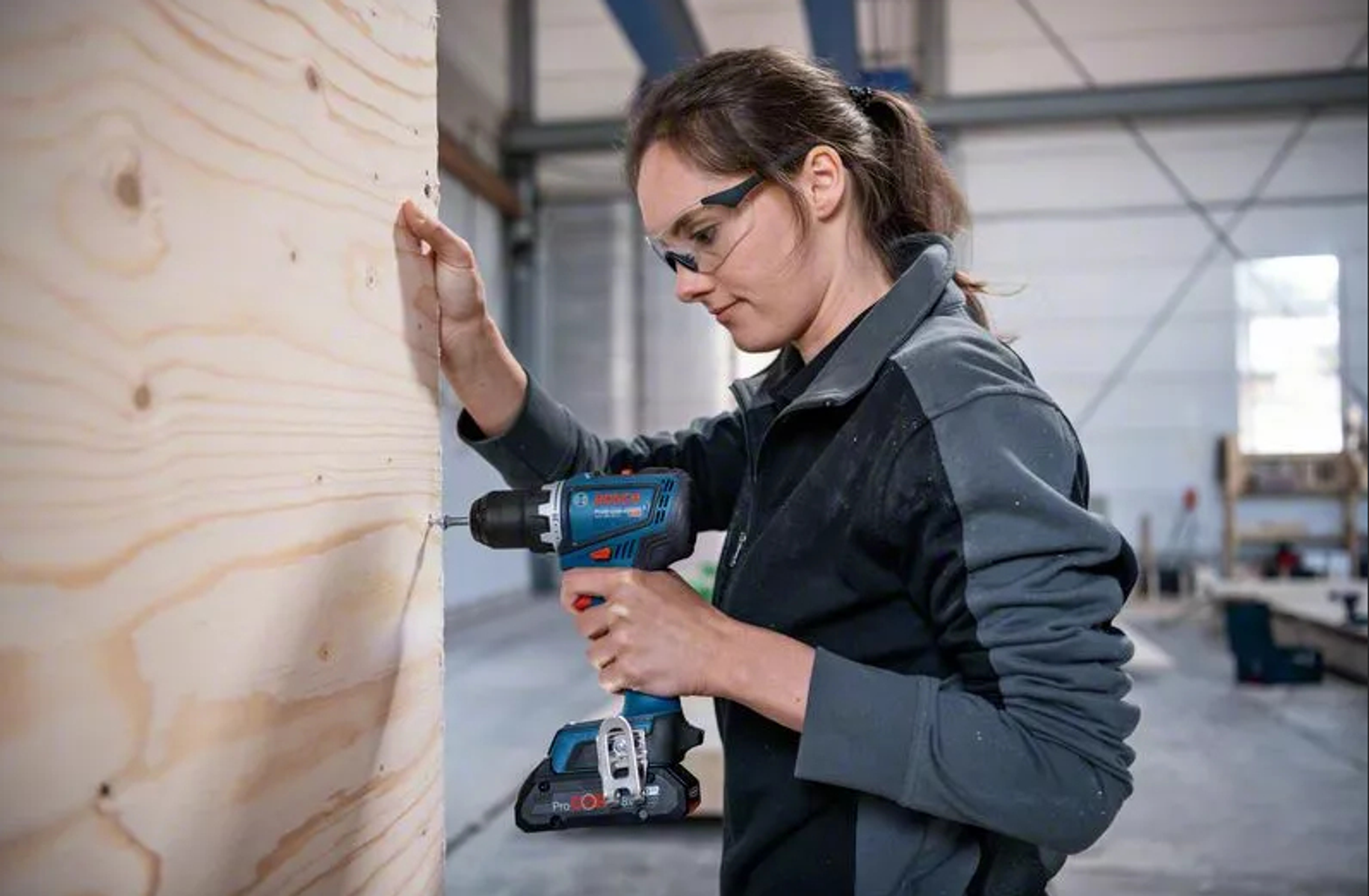 Kobieta wierci w drewnie niebiesko-czarną wiertarką, nosząc okulary ochronne.