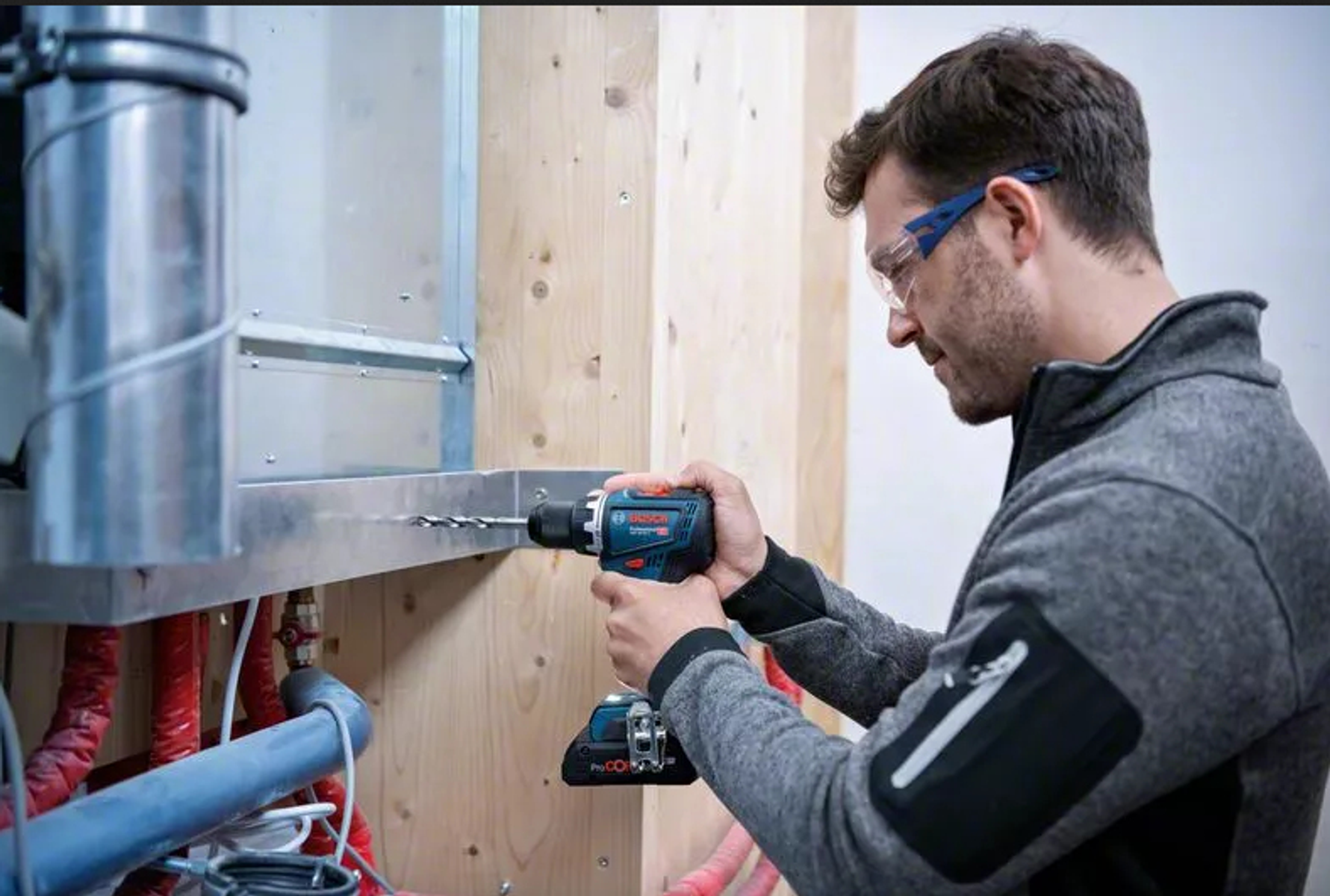 Mężczyzna wierci w drewnie. Ma okulary ochronne. Używa wiertarki. Szary sweter. Wnętrze.