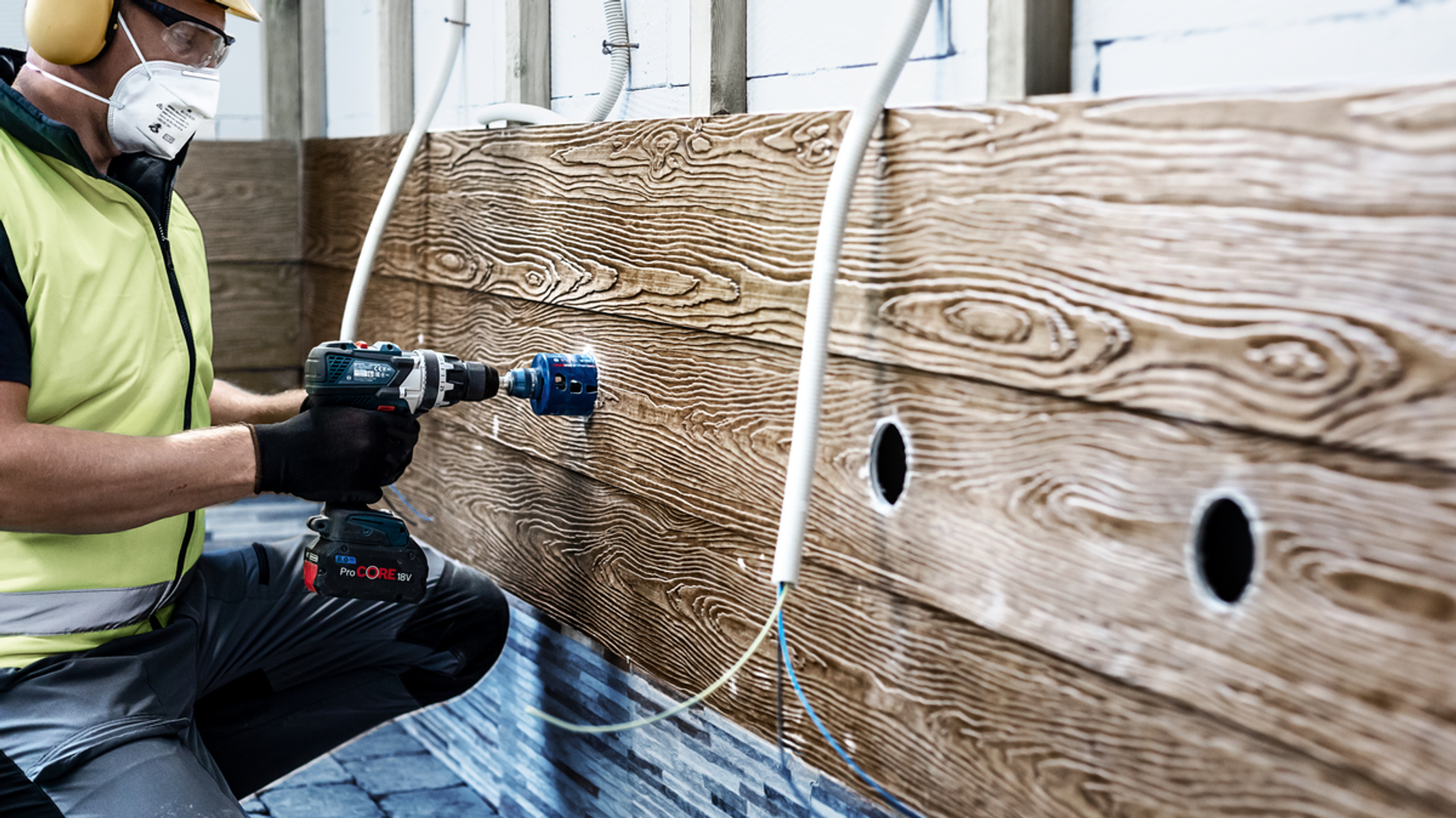 Osoba używająca wiertarki do wywiercenia otworu w drewnianej ścianie z rurami w tle.