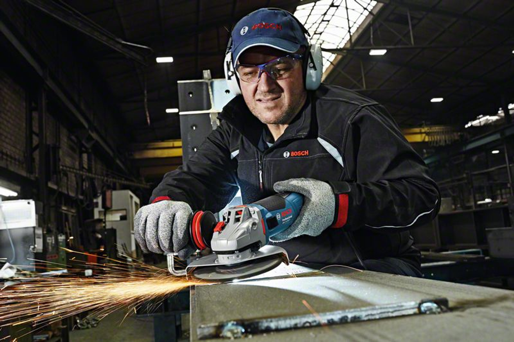Mann schleift Metall mit Winkelschleifer, Funken fliegen, trägt Schutzkleidung, in einer Werkstatt.
