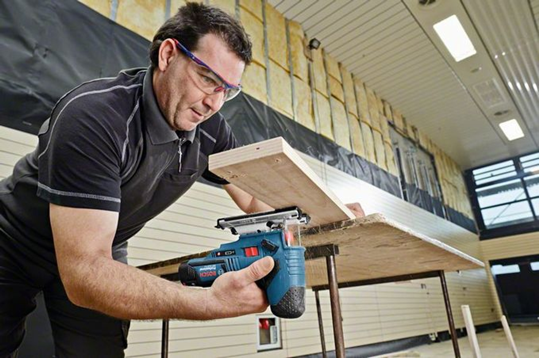 Mężczyzna używający wyrzynarki do cięcia drewnianej deski, w okularach ochronnych.