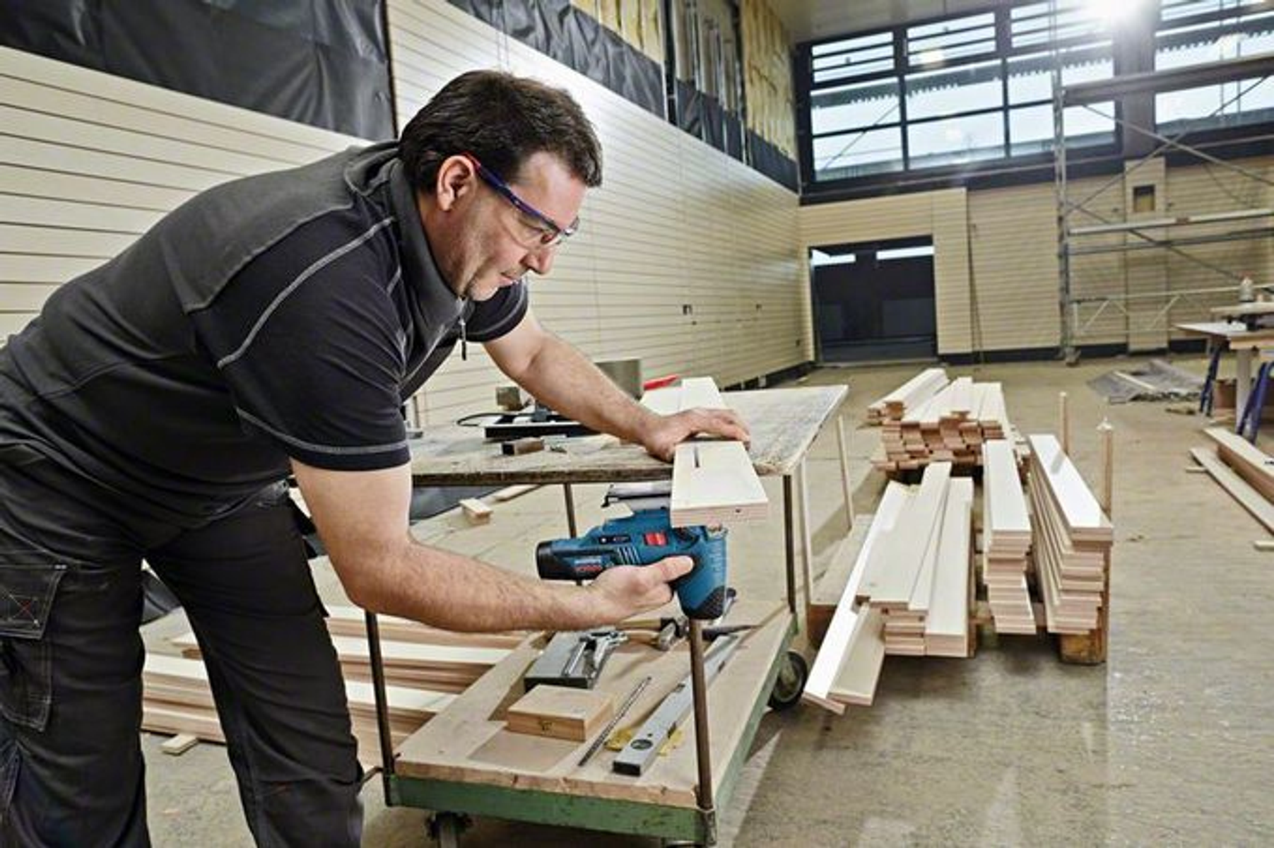 Mężczyzna używa niebieskiej wyrzynarki do drewna, warsztat, widoczne narzędzia.