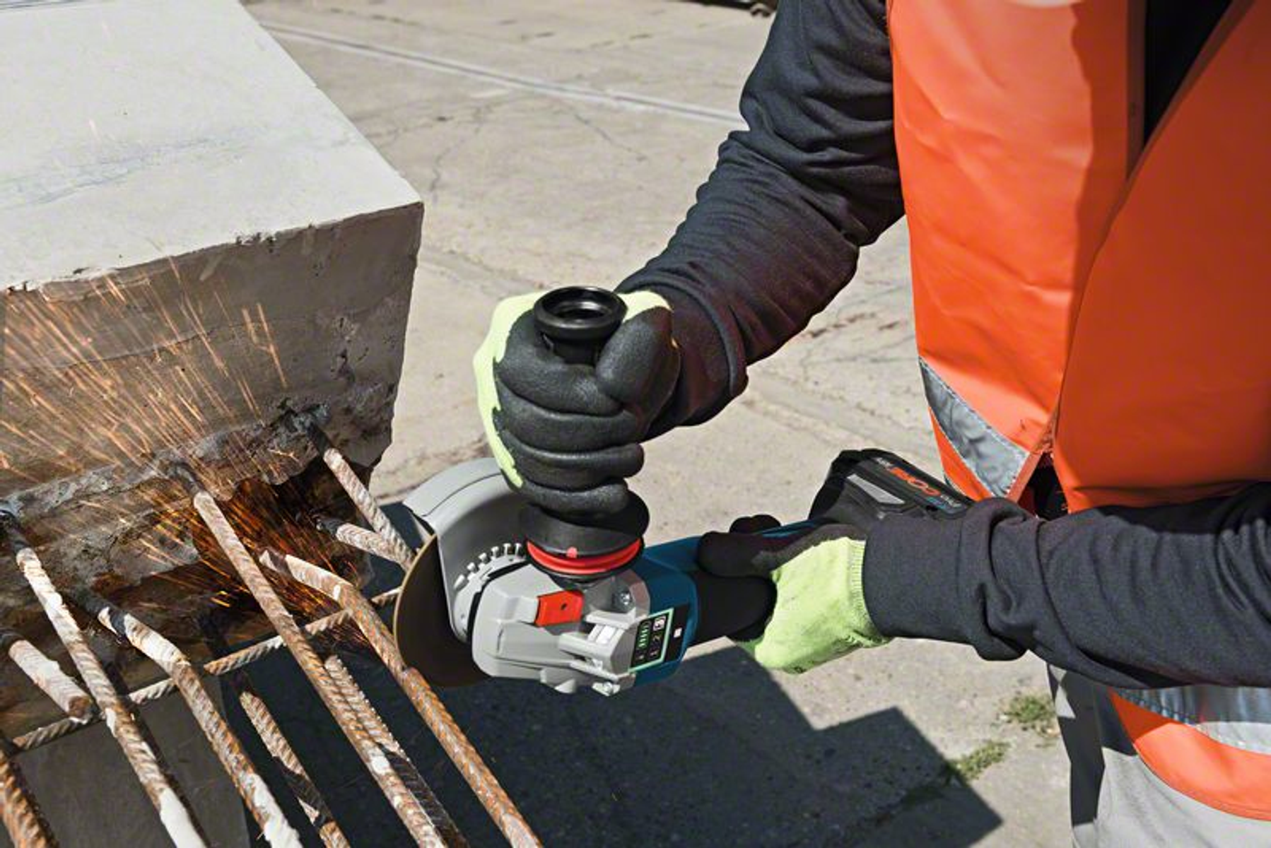 Robotnik używający szlifierki kątowej, przecina pręt zbrojeniowy. Widoczna kamizelka i rękawice ochronne.