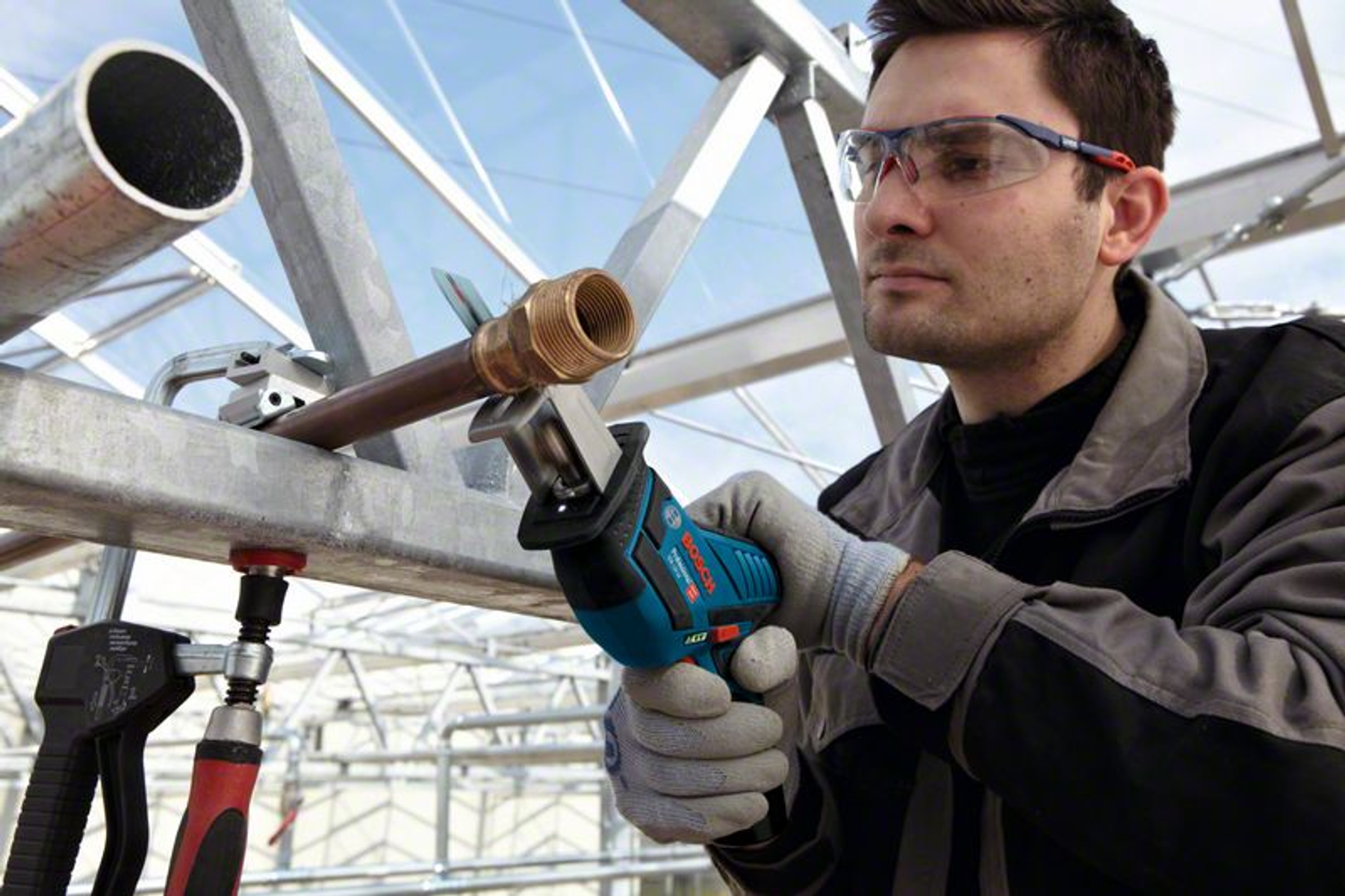 Mężczyzna używający piły szablastej Bosch. Ma na sobie okulary ochronne i rękawice. Szklarnia w tle.