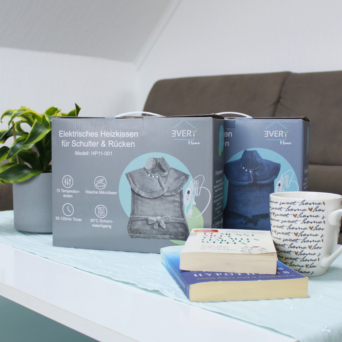 Zwei Kartons eines elektrischen Heizkissens, Bücher und eine Kaffeetasse auf einem Tisch.