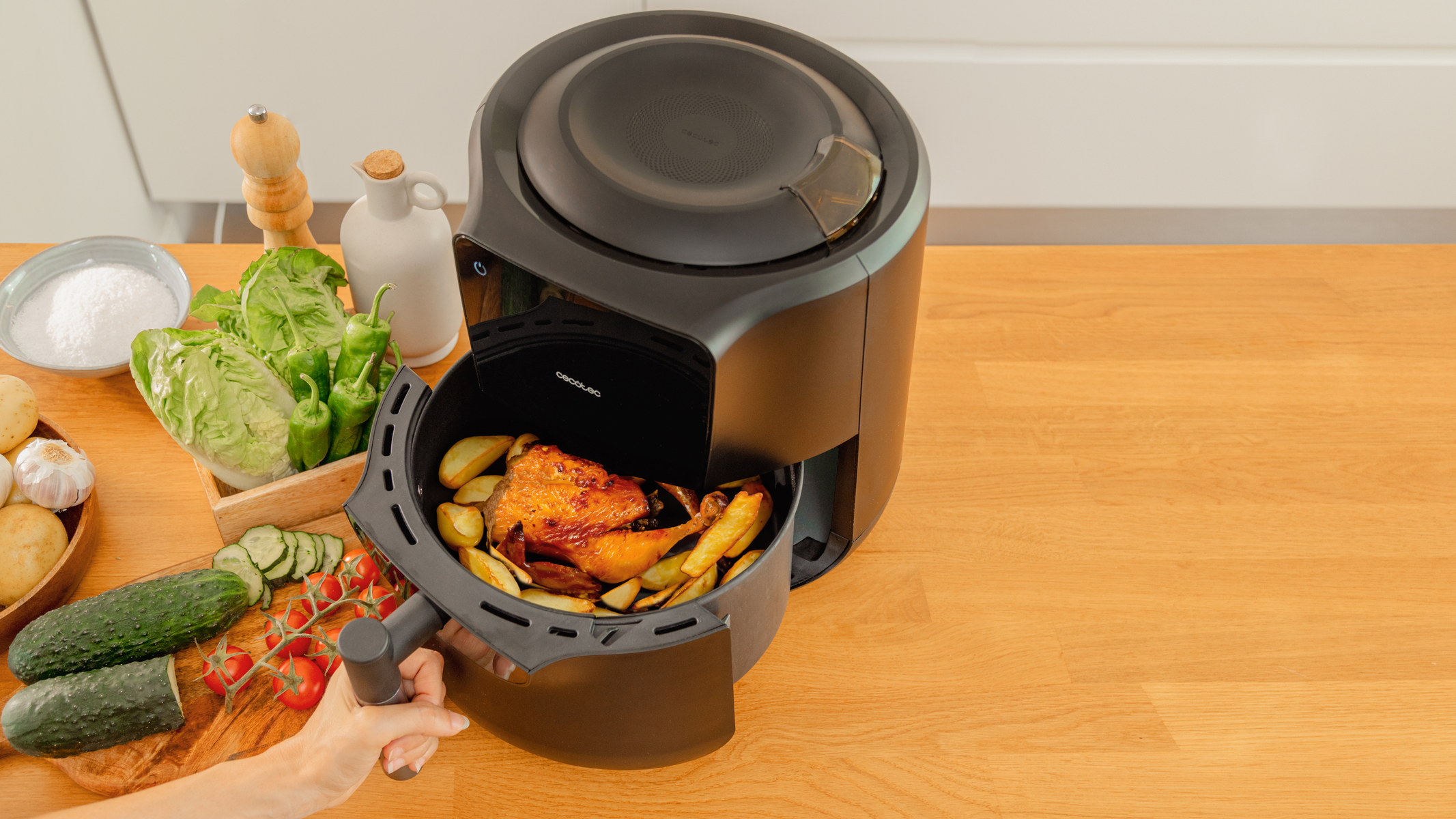 Una freidora de aire con pollo y patatas dentro. Verduras y condimentos sobre una superficie de madera.