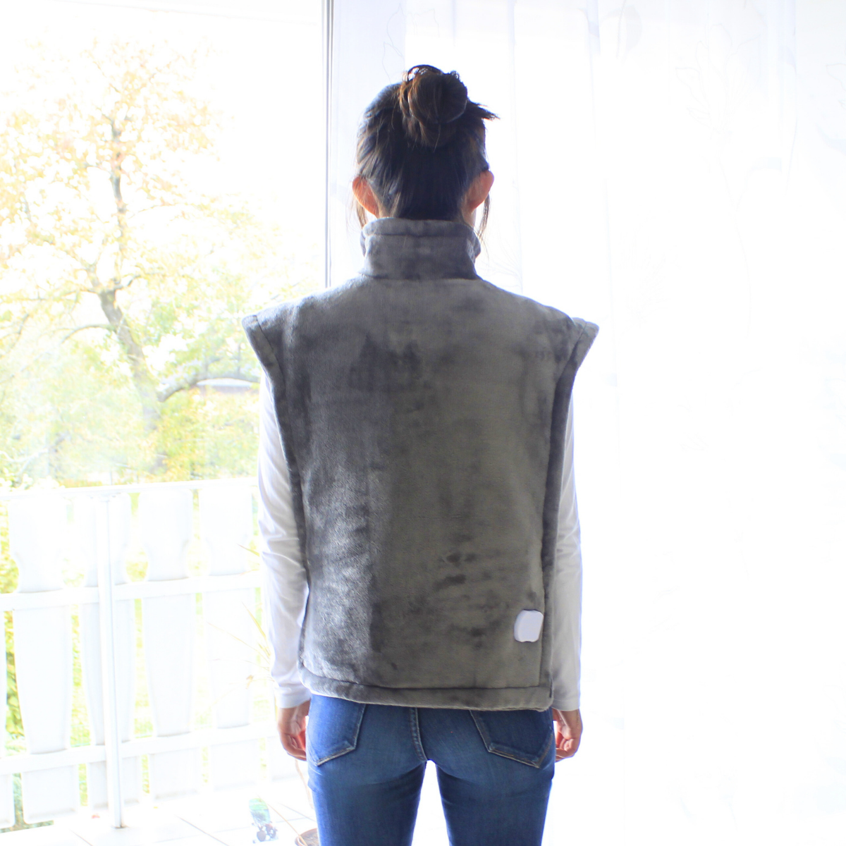 Eine Frau von hinten mit einer grauen beheizten Weste. Sie steht vor einem Fenster mit Aussicht.