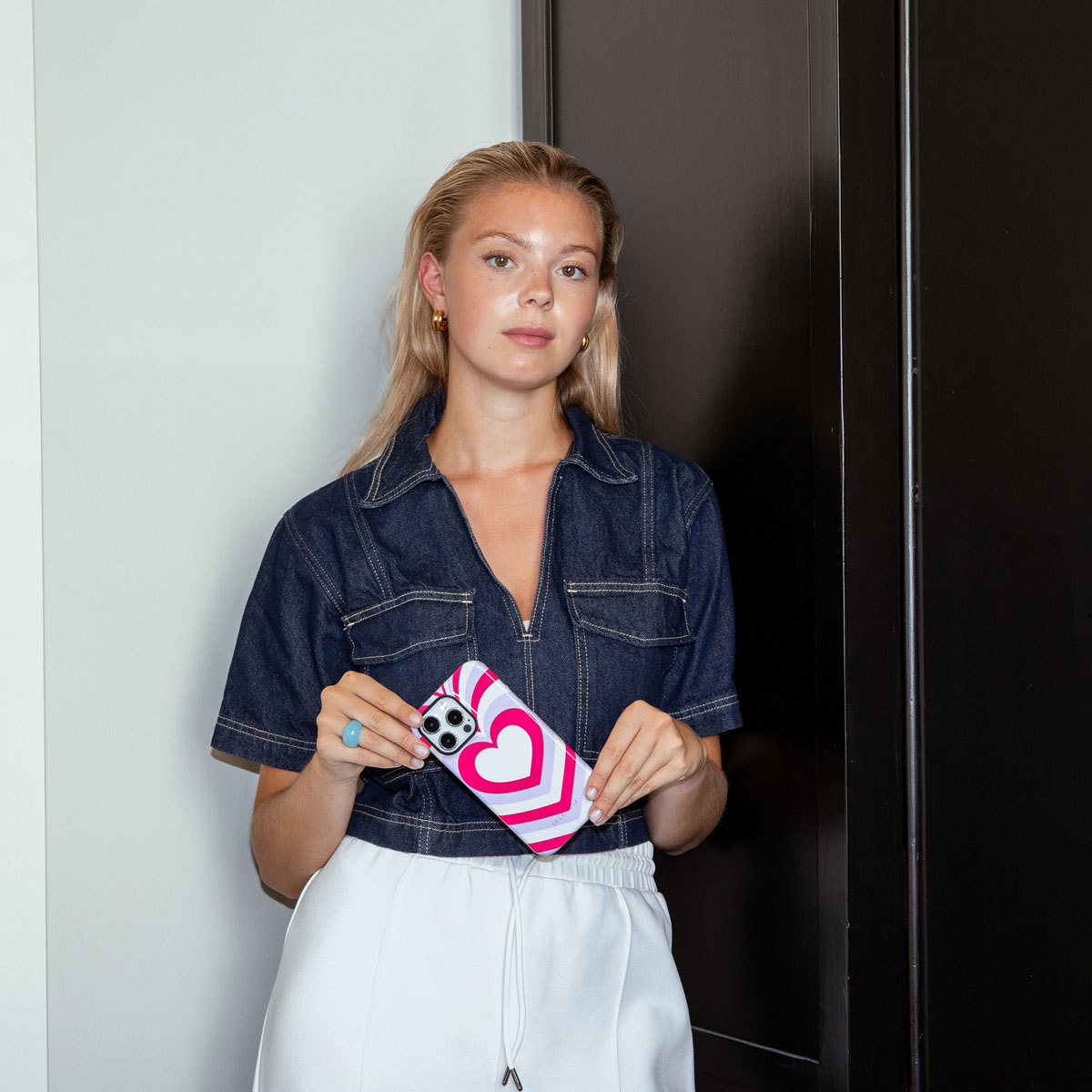 Une femme tient un téléphone avec un motif de cœur rose. Elle porte une chemise en jean et un pantalon blanc.