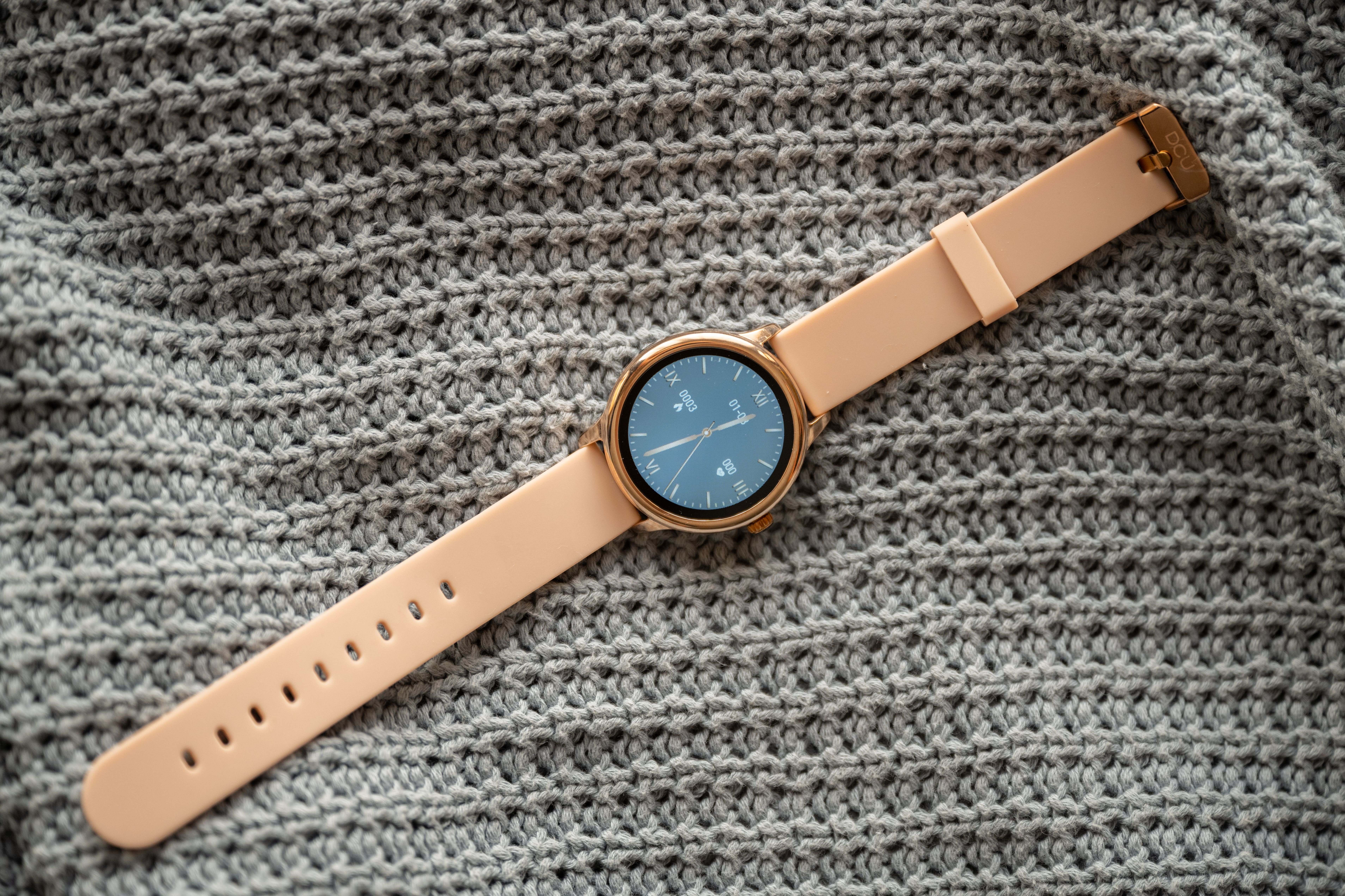 Złoty i brzoskwiniowy smartwatch na szarej dzianinie.