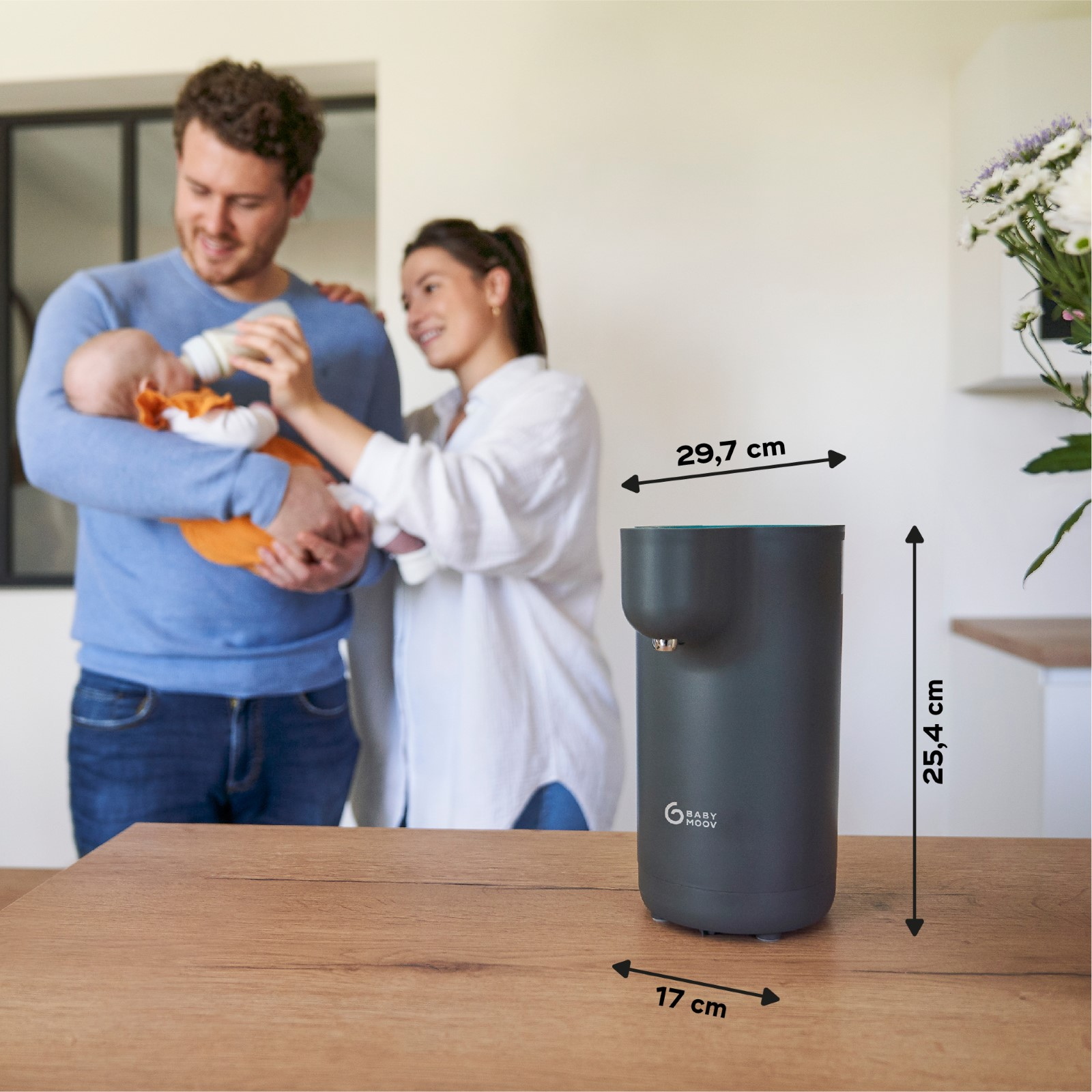 Familie füttert Baby mit Flasche. Dunkelgrauer Babyflaschenwärmer auf Holztisch, mit Abmessungen.