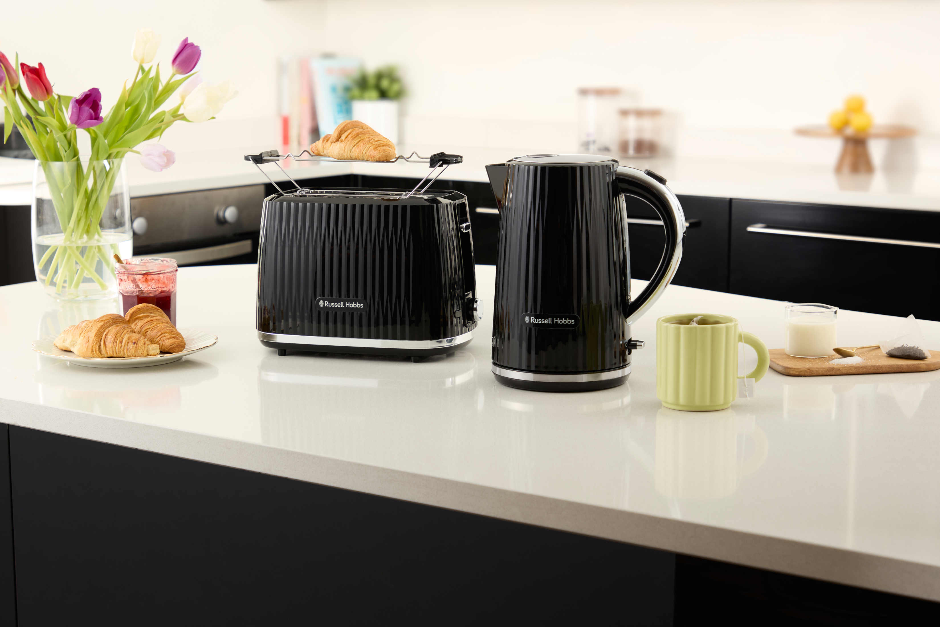 Czarny toster i czajnik na białym blacie z rogalikami i kubkiem.