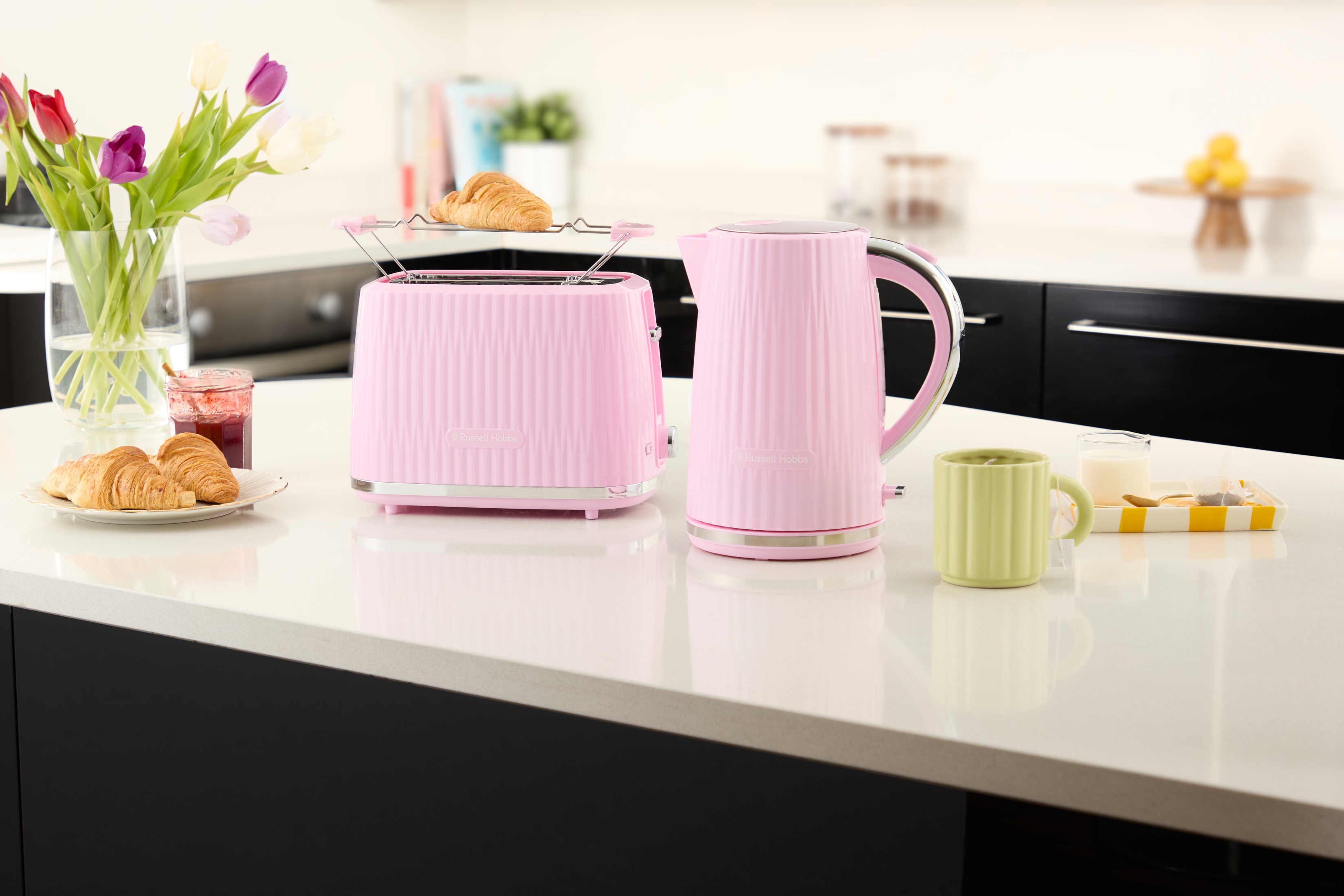 Różowy toster, czajnik i kubek na białym blacie. Kwiaty i rogaliki z boku. Scena śniadaniowa.