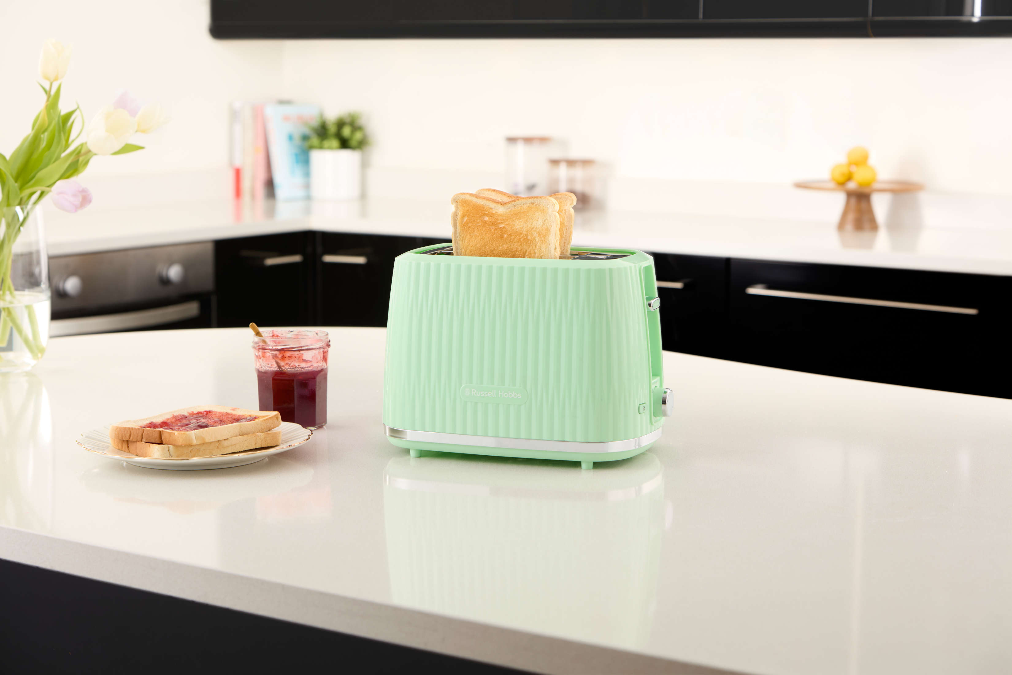 Miętowo-zielony toster z chlebem w środku, talerz z chlebem i dżemem, tulipany w wazonie, na białym blacie.