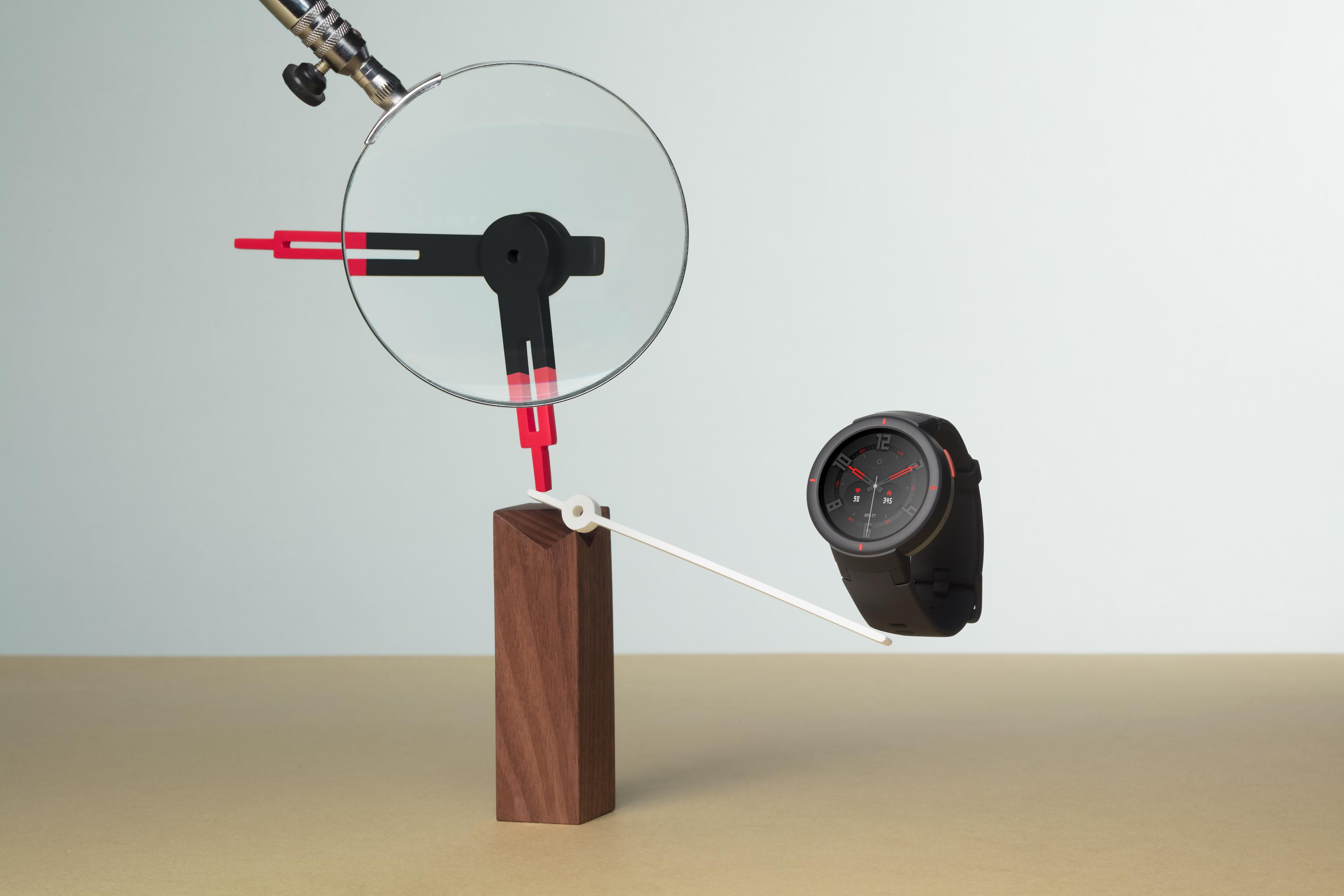 Un reloj con detalles negros y rojos flota en el aire, con una lupa encima, sobre una superficie beige claro.