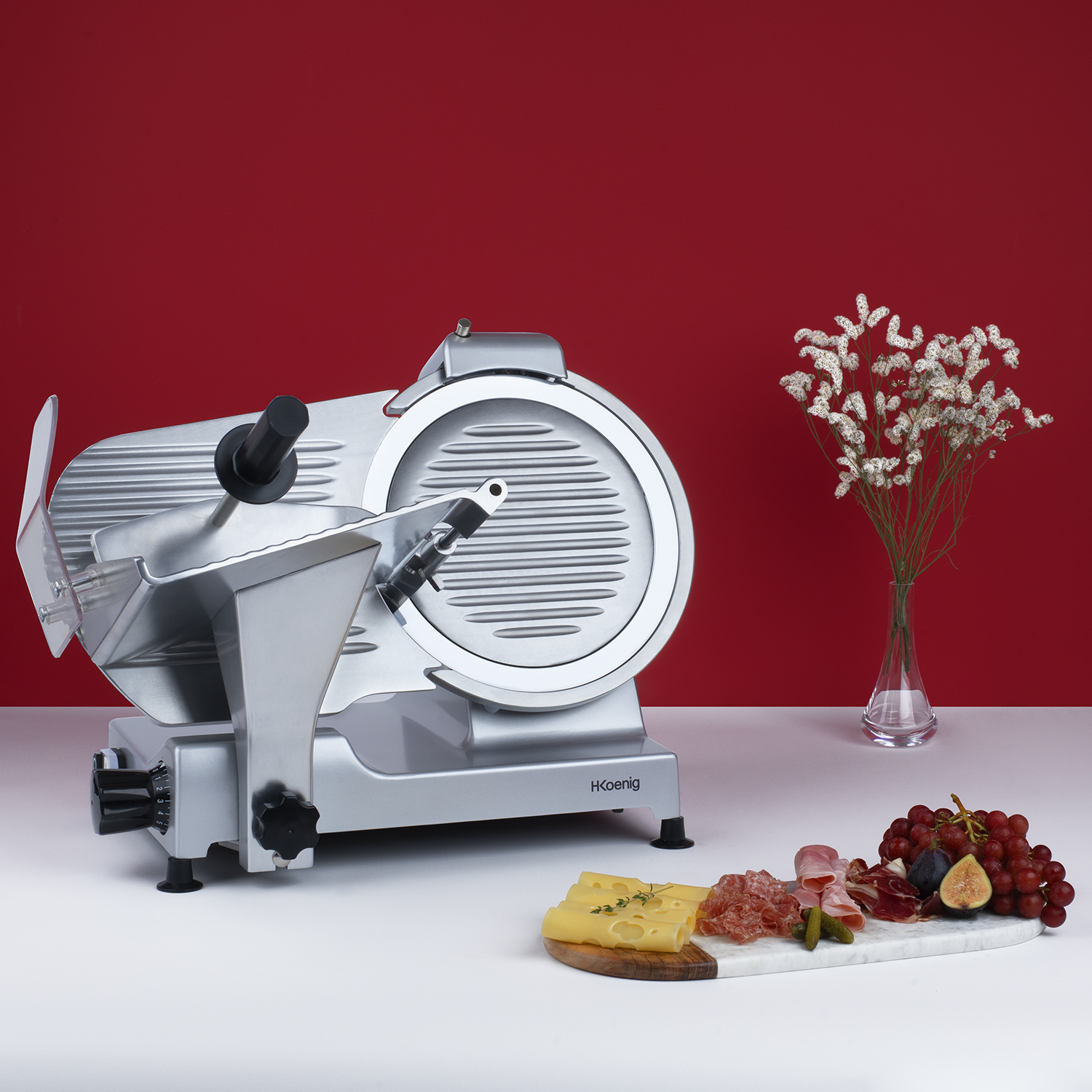 Srebrna krajalnica do mięsa na białej powierzchni, z serem, salami, figami i winogronami. Czerwone tło.