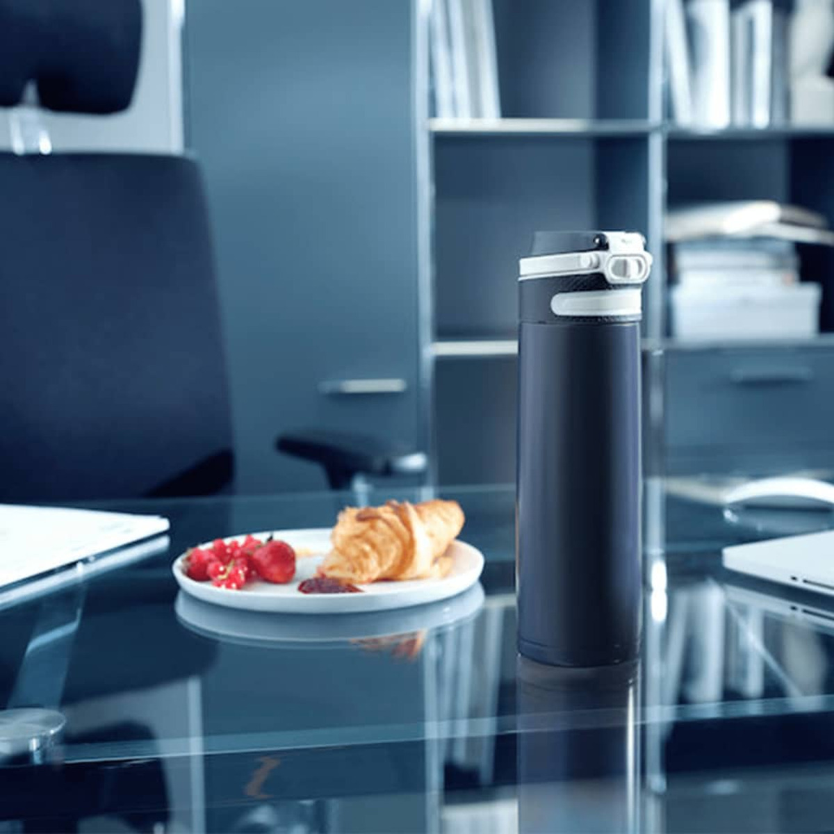Eine Thermoskanne aus Edelstahl auf einem Glastisch mit einem Teller mit Essen und einem Laptop im Hintergrund.