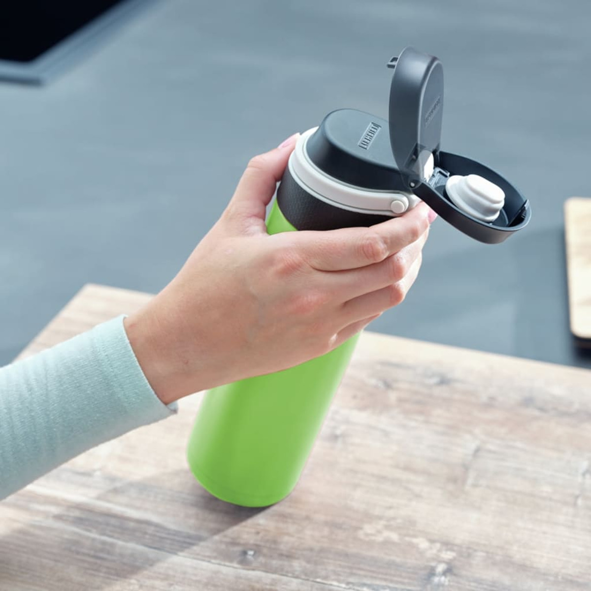 Hand hält eine grün-schwarze Wasserflasche mit geöffnetem schwarzem Deckel. Hintergrund unscharf.