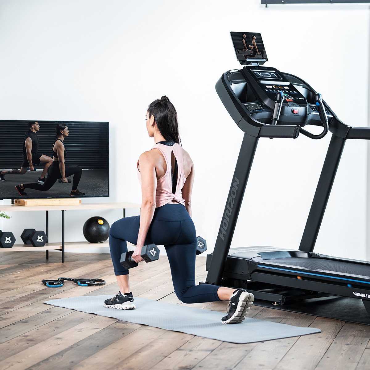 Kobieta robi wykroki z hantlami, przed bieżnią i TV z ćwiczeniami.